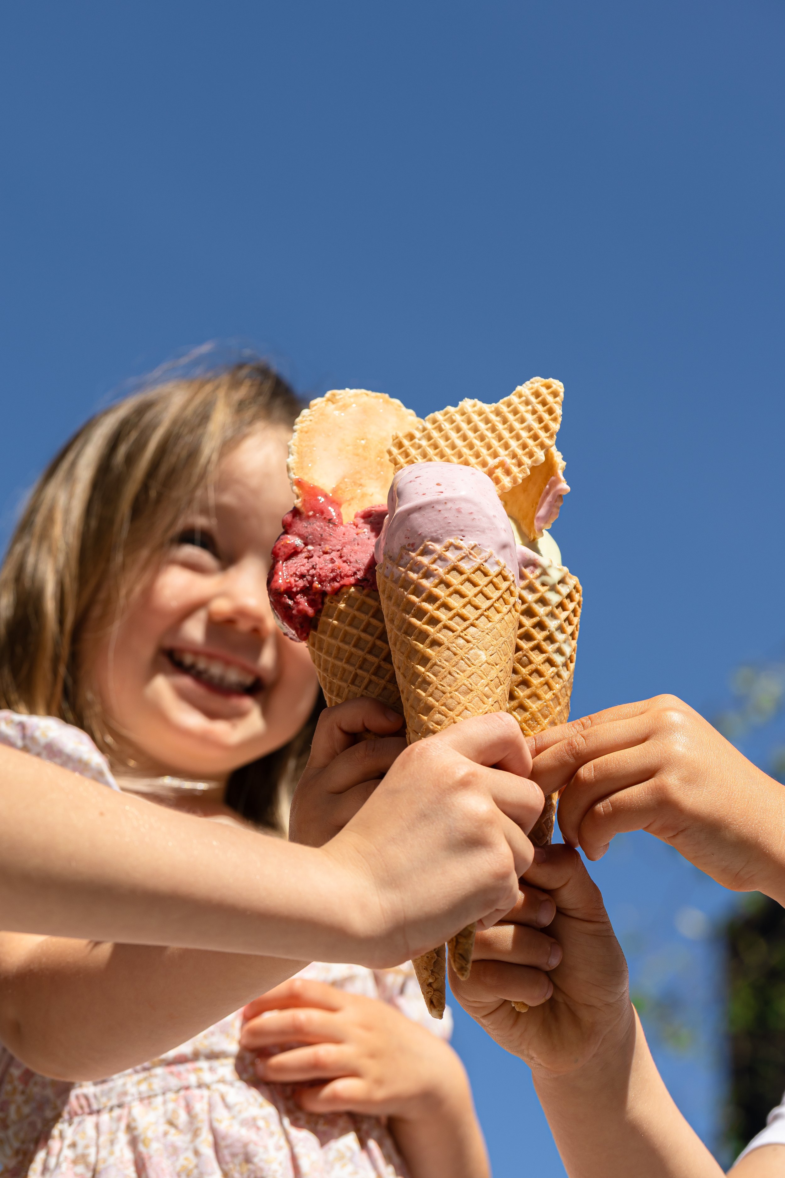 Children holding a large ice cream cone with multiple scoops and waffle cookies, smiling against a bright blue sky.