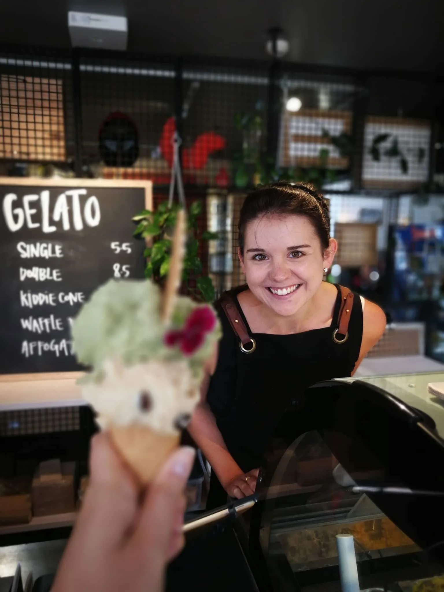 Smiling woman behind a counter in a gelato shop, menu board listing various gelato flavors, some plants and decorative items in the background.