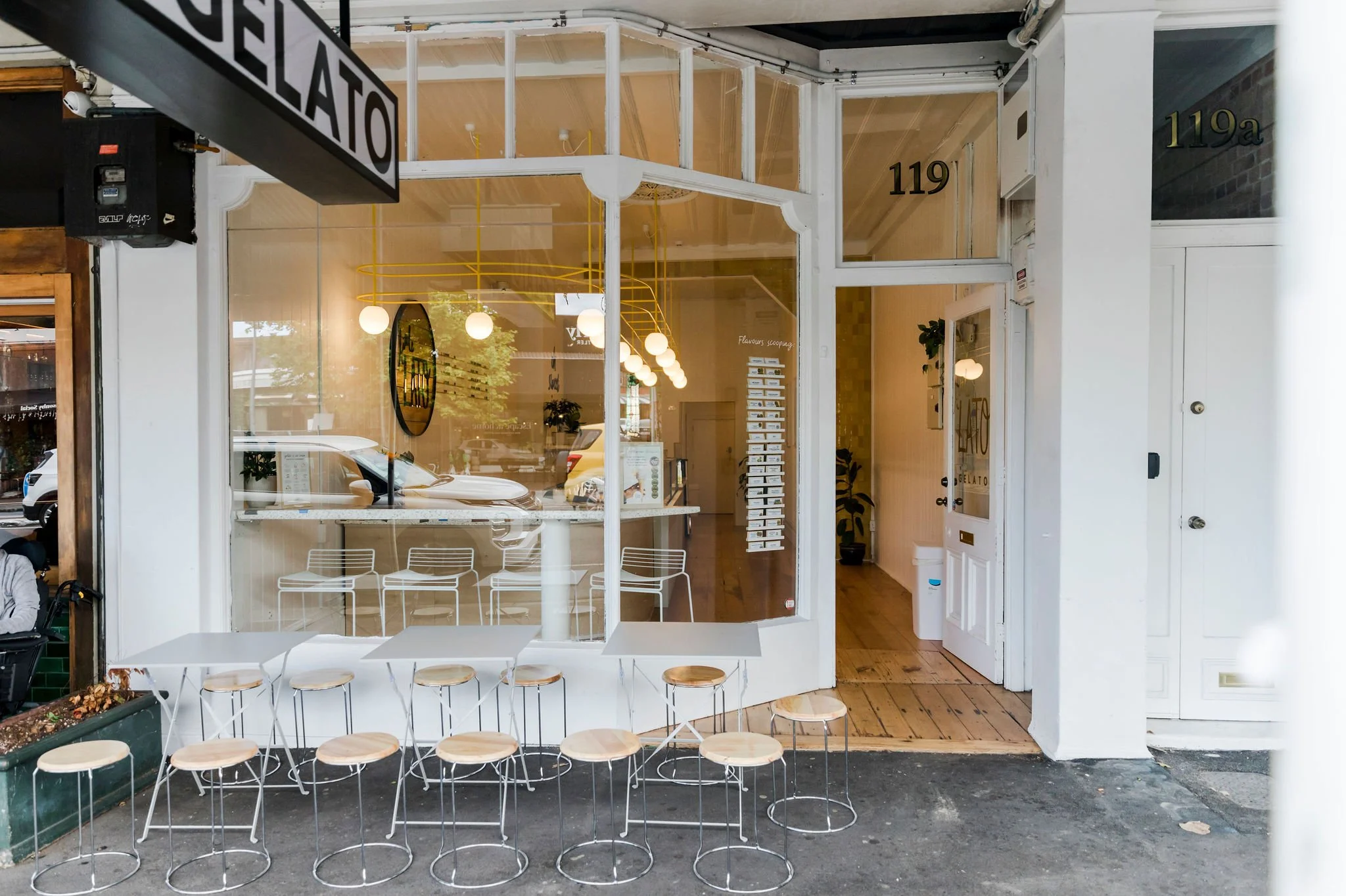Exterior of a small café or gelato shop with a large glass window and white frame, outdoor seating with round wooden tables and stools. A sign that reads 'GELATO' is partially visible hanging above. The interior features modern lighting and decor, with a view of chairs and a counter inside.