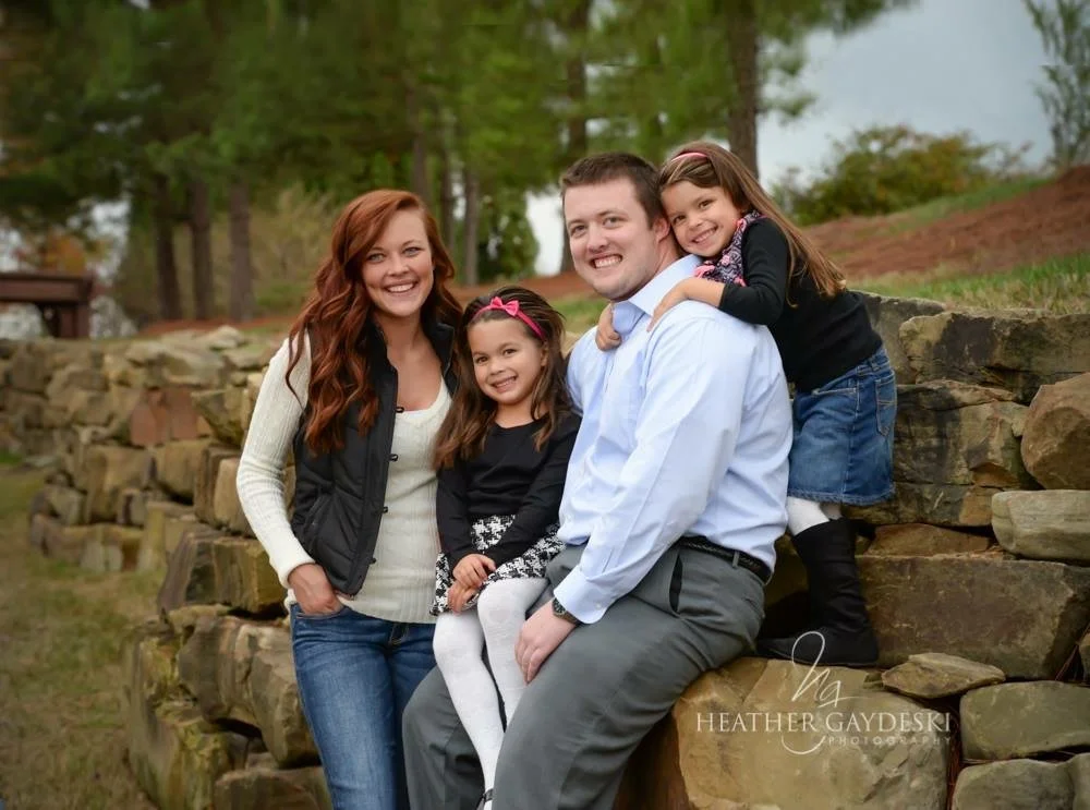 Cristina Moore's Family sitting on rocks outdoors, smiling at the camera. Trees and greenery are in the background.