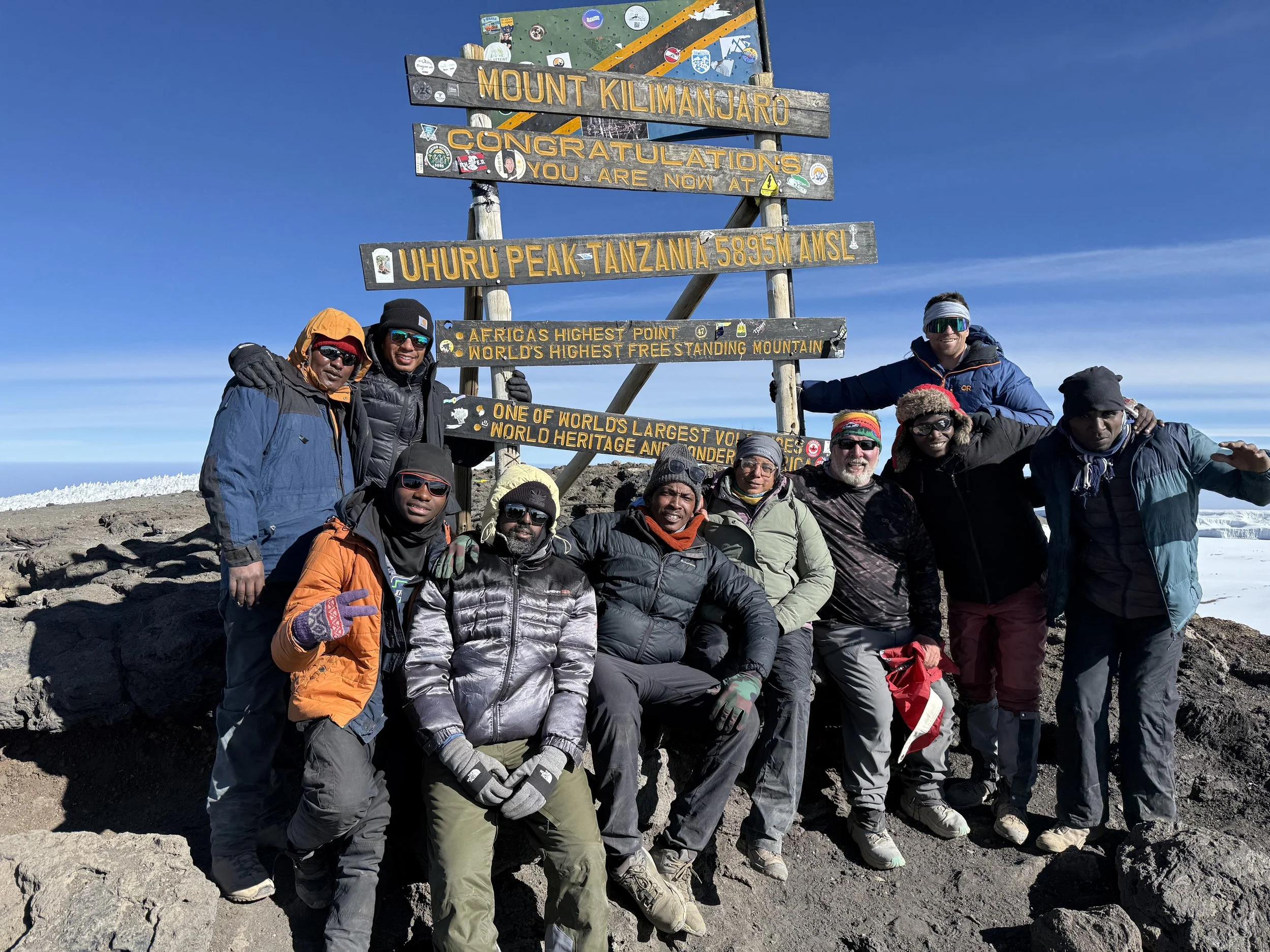 Cristina Moore with group climbers at Uhuru Peak on Mount Kilimanjaro in Tanzania, posing under a sign that marks the summit at 5895 meters above sea level. The sky is clear and the terrain is rocky with snow in the background.