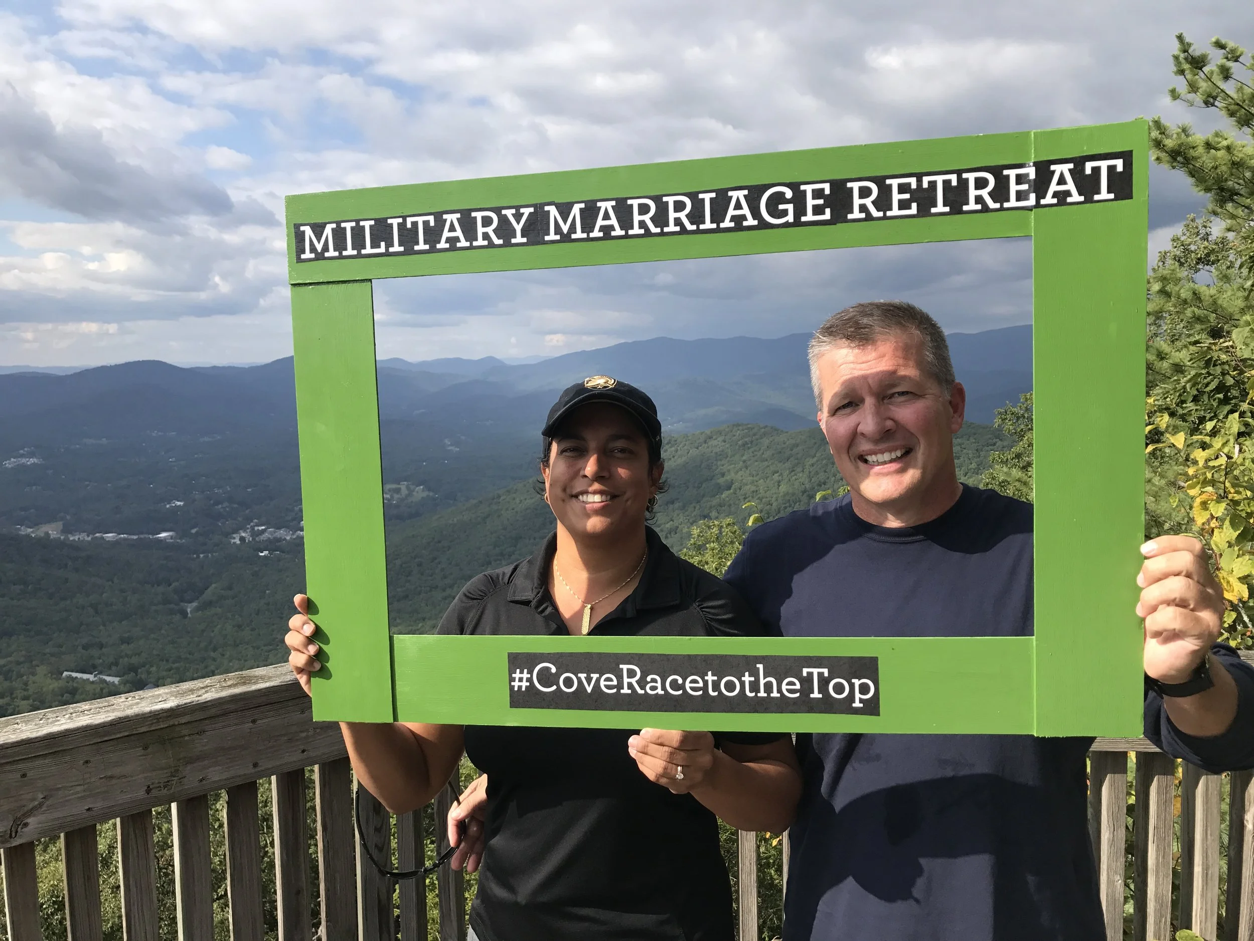 Cristina Moore and her husband holding a green frame sign that reads 'Military Marriage Retreat' and '#CoveRacetoTheTop' with a mountain landscape in the background.