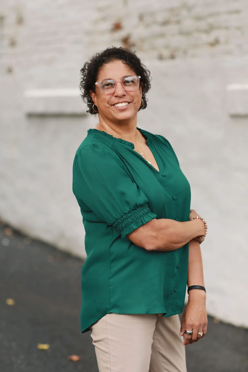 Cristina Moore, a Christian Author, smiling with arms crossed, wearing a green blouse and beige pants, standing outdoors against a light-colored brick wall.