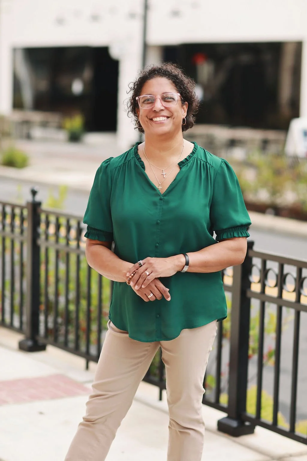 Cristina Moore, a faith-based author, smiling outdoors, wearing glasses, a green blouse, and beige pants, standing near a black metal fence with a blurred urban background.