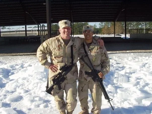 Cristina Moore, a Christian writer and military vet in camouflage uniforms holding rifles, standing side by side outdoors on snow with a covered structure and trees in the background.