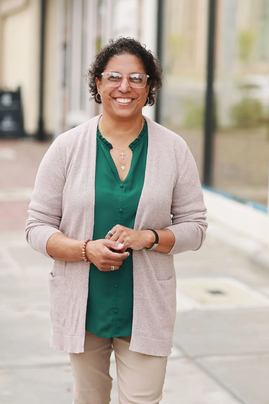 Cristina Moore, a Christian speaker and writer, standing outdoors near glass windows, wearing a green blouse and beige cardigan, accessorized with necklaces and bracelets.