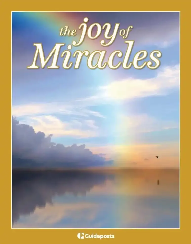 A beach scene with a rainbow stretching into the sky, clouds, and a calm body of water reflecting the sky. Text overlay says 'the joy of Miracles'.