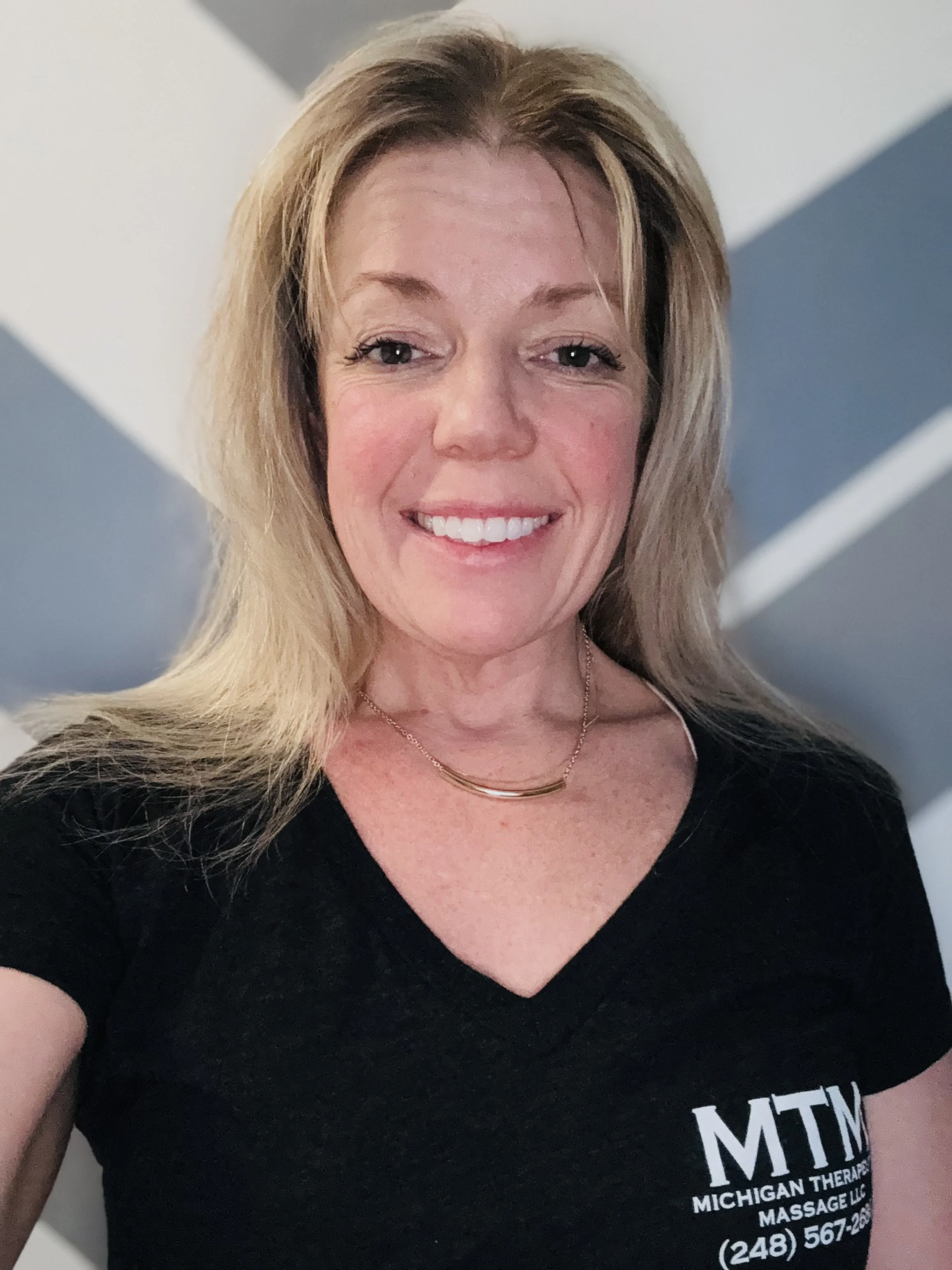 A smiling woman with blonde hair, wearing a black T-shirt with white text and a gold necklace, standing in front of a wall with gray and white geometric patterns.