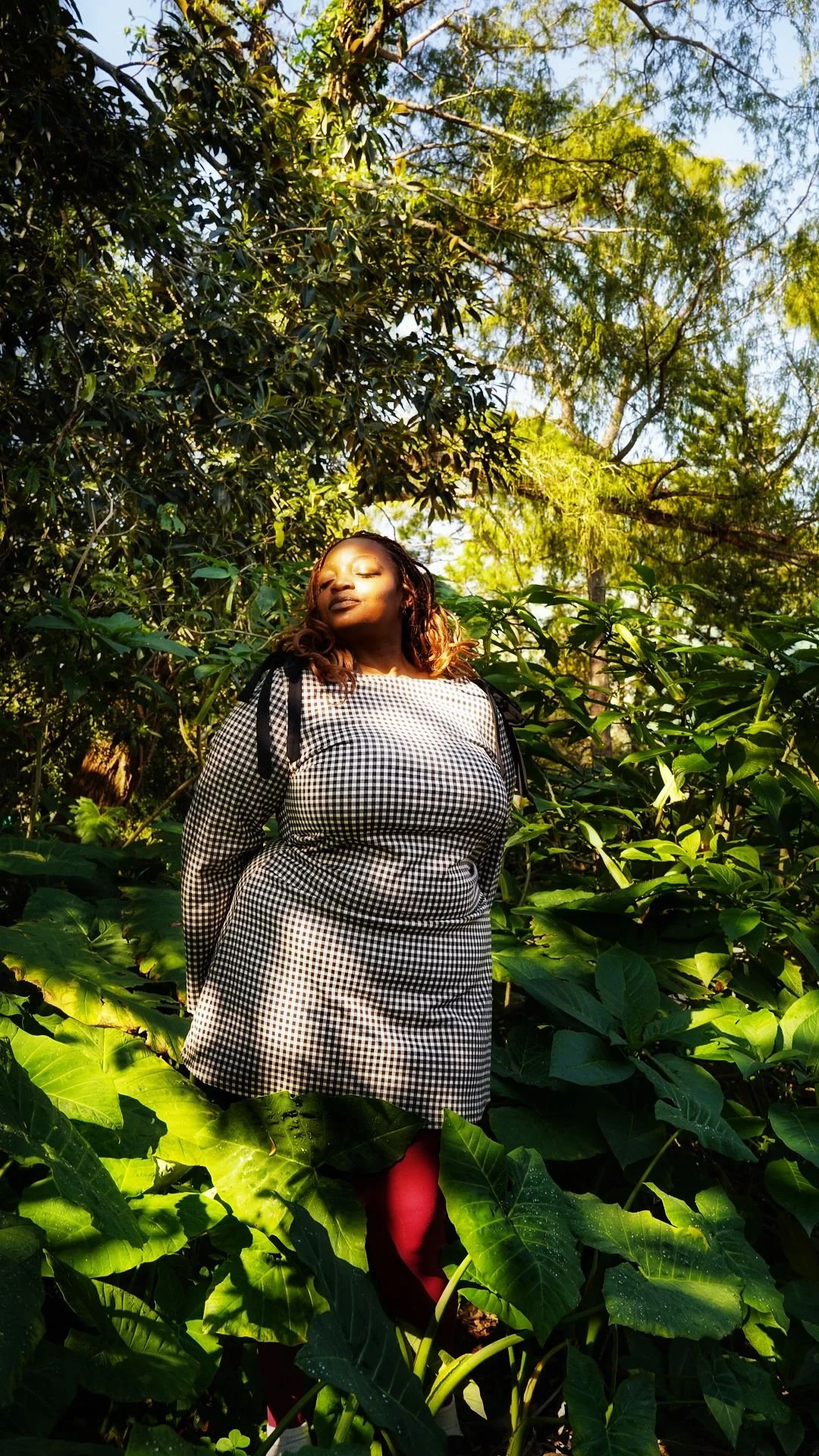 A woman with braided hair is standing among large green leaves outdoors, facing the sunlight with her eyes closed and a peaceful expression.