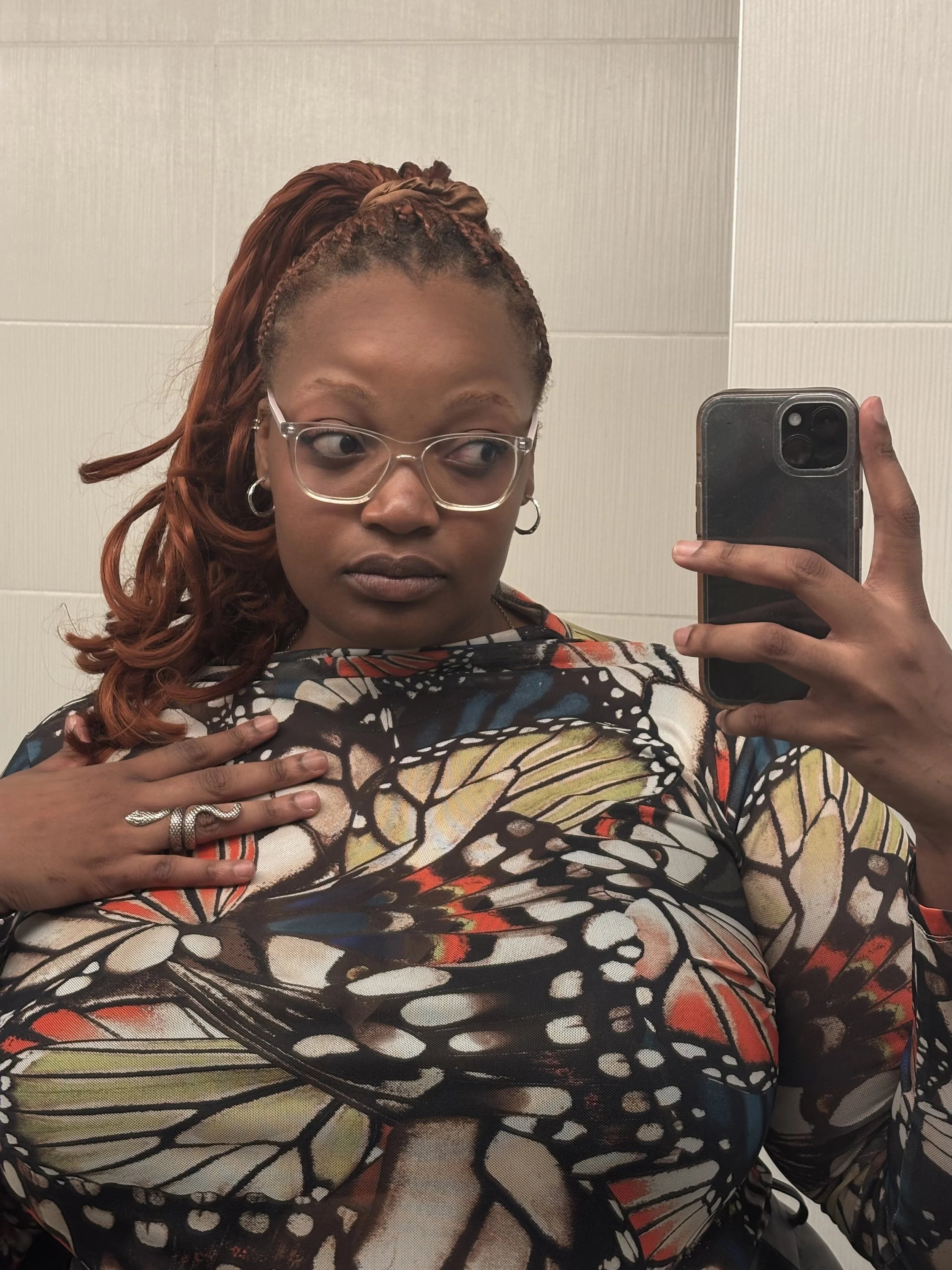 Woman with glasses taking a selfie in front of a mirror, wearing a colorful butterfly-patterned top, with her hand on her chest and hair styled in a side ponytail.