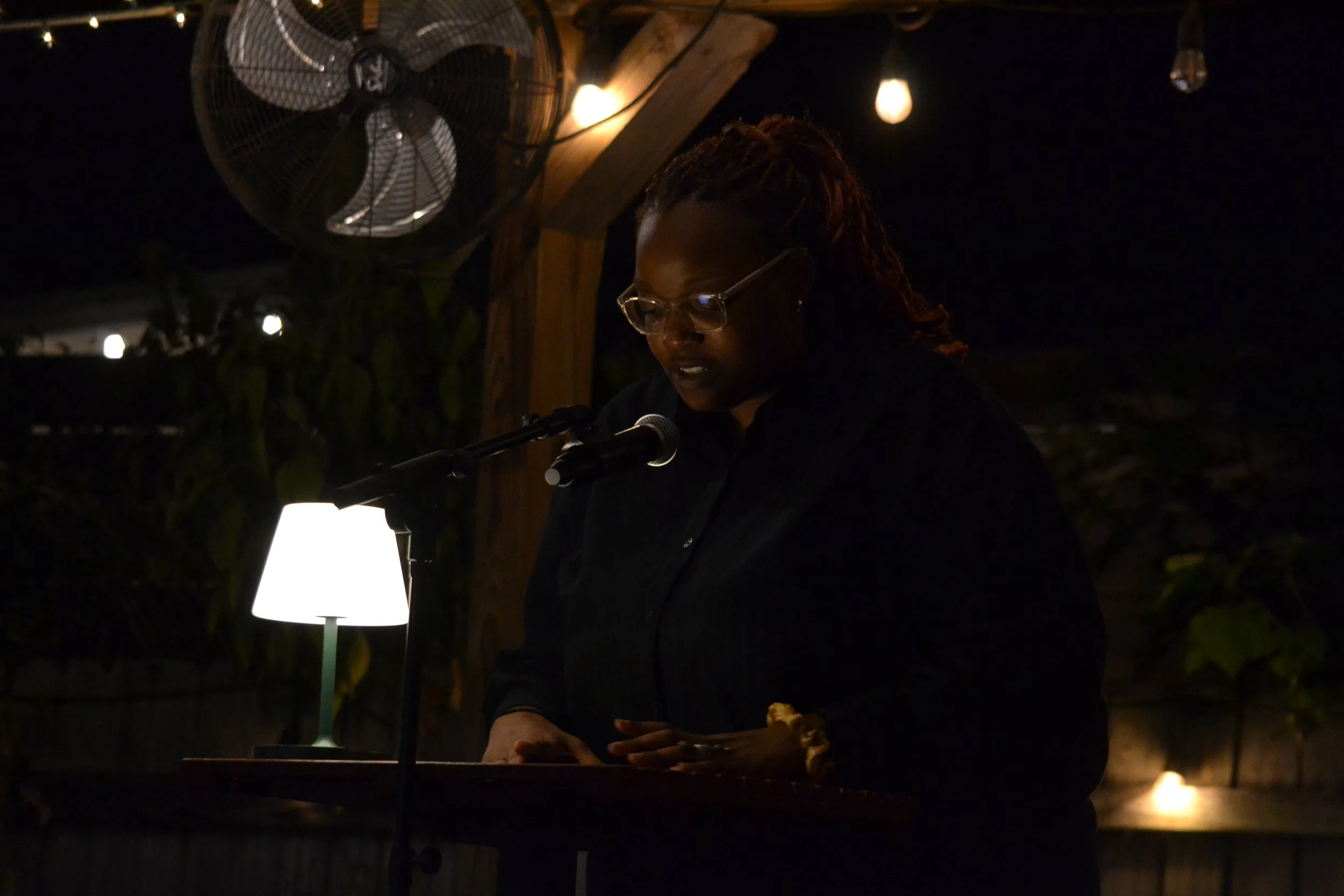 A woman with glasses and braided hair reading or speaking at a podium with a microphone in an outdoor setting at night, illuminated by a small table lamp and string lights.