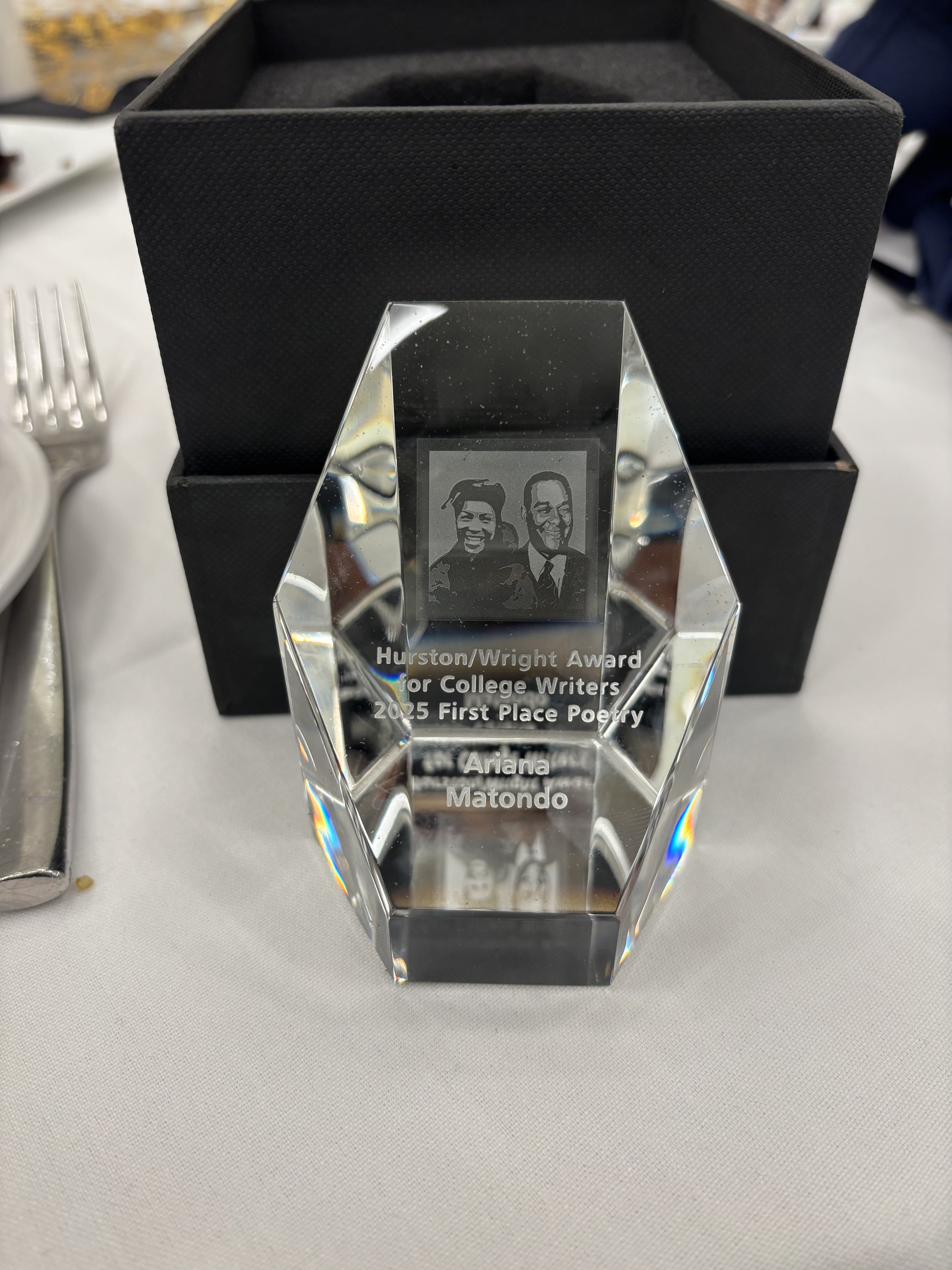 Clear crystal award with an etched image of two smiling men, inscribed with 'Hurston/Wright Award for College Writers 2025 First Place Poetry, by Ariana Matondo,' placed on a white tablecloth with a black box behind it.