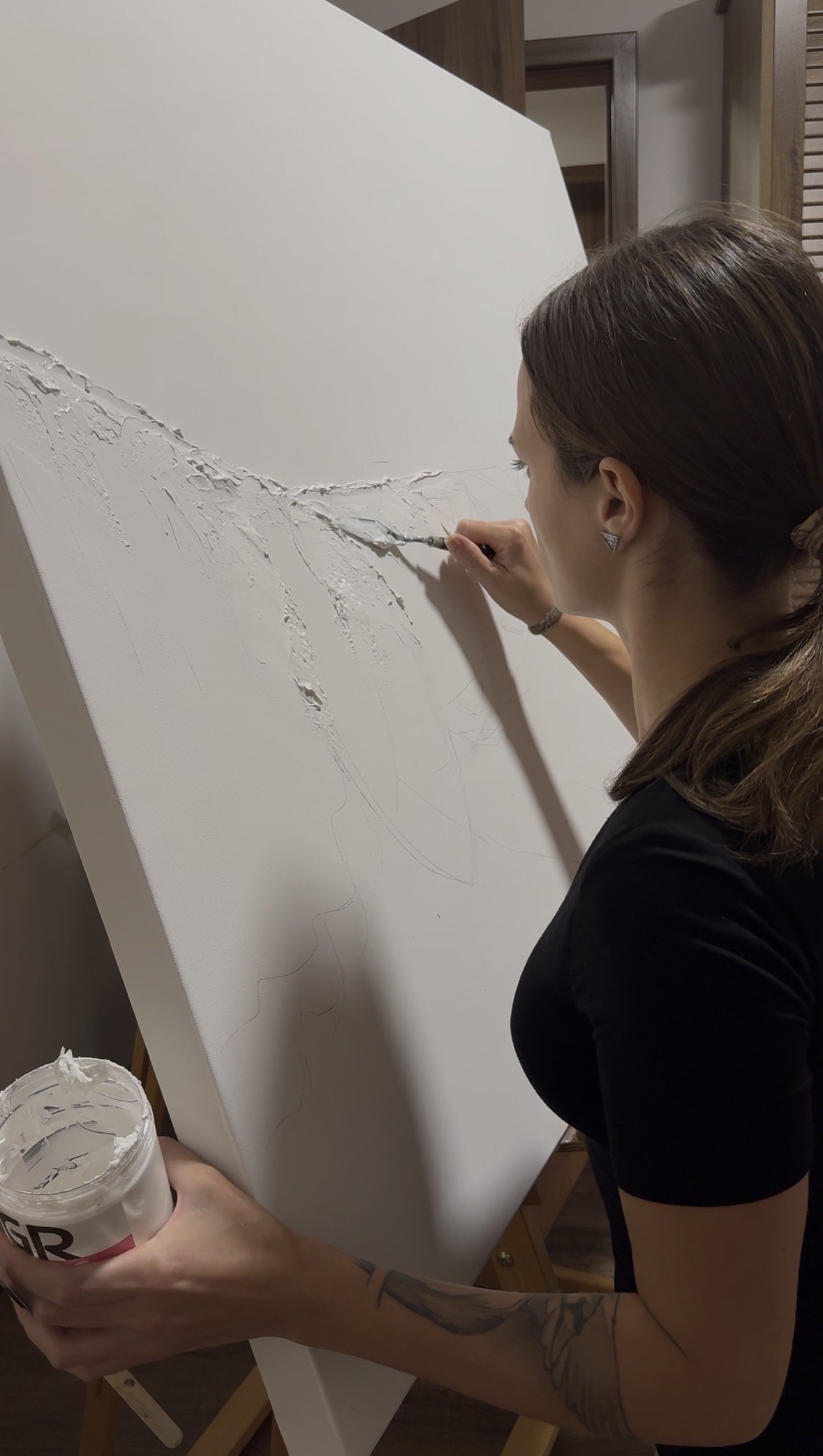 Woman painting a textured white abstract art piece with a small brush while holding a container of white paint, standing in a room with wooden accents.