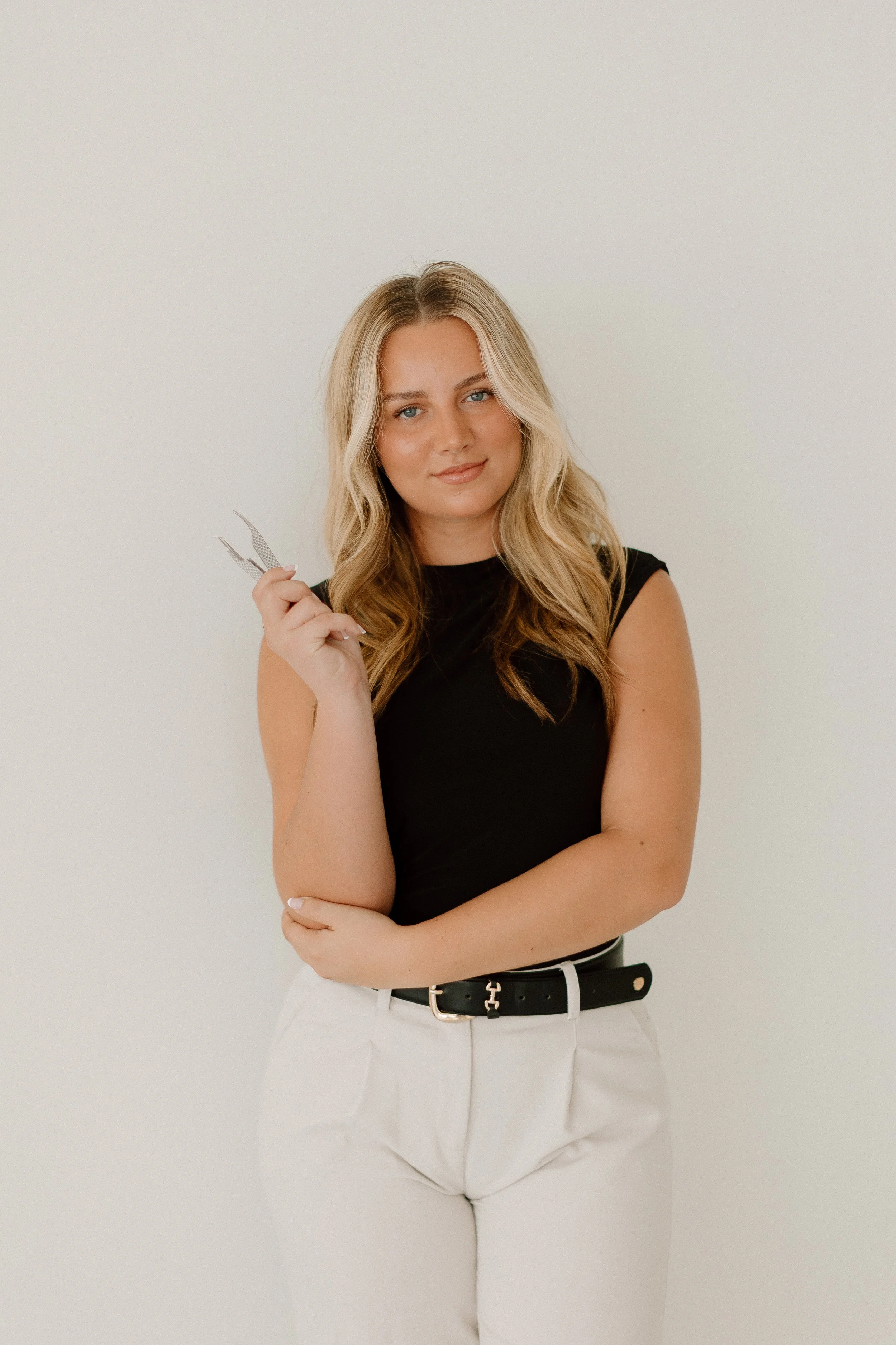 A woman with wavy blonde hair, wearing a black sleeveless top and cream-colored pants, stands against a plain off-white background holding a pair of tweezers in her right hand.