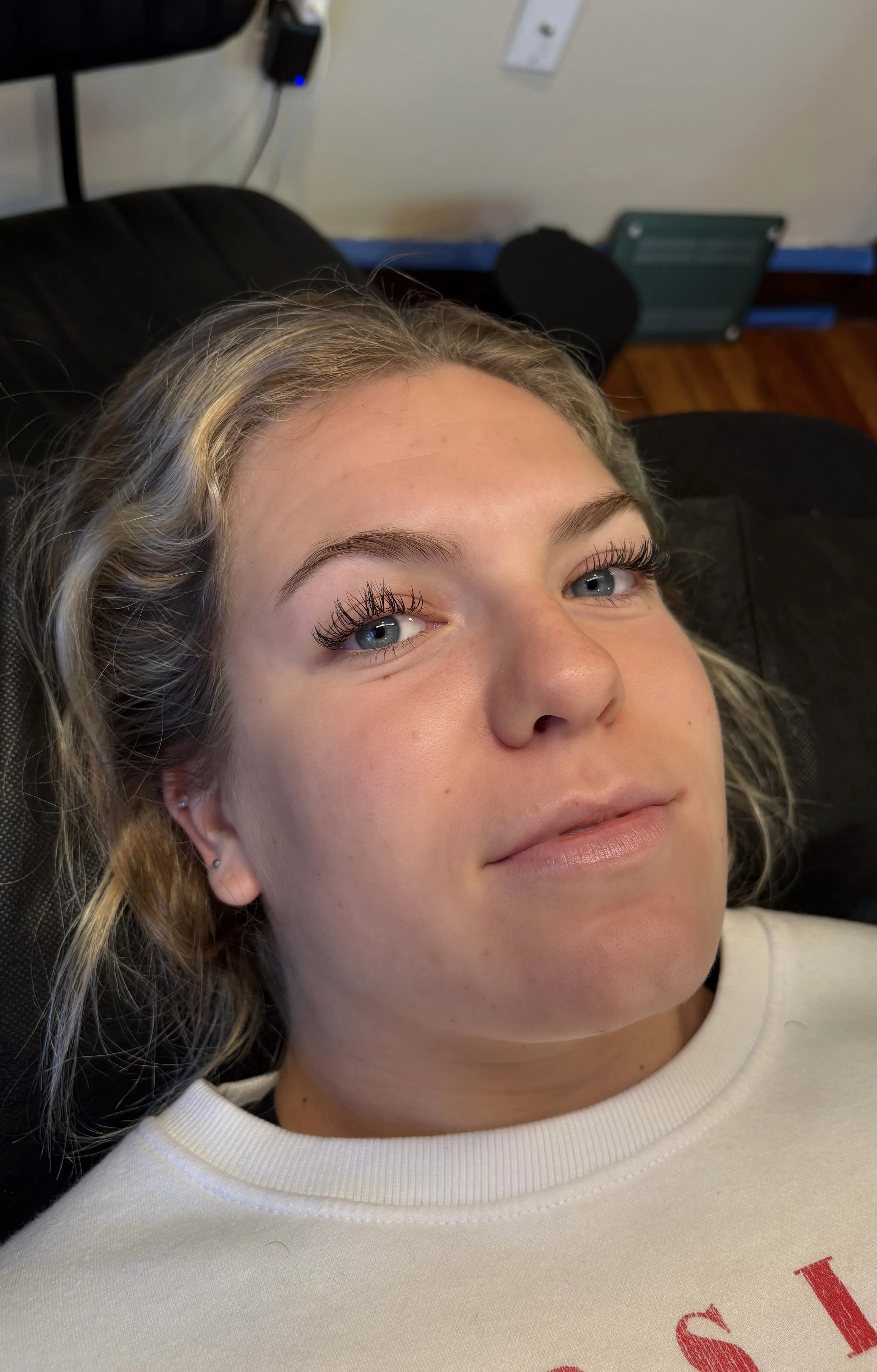 Close-up of a woman lying on a black surface with a relaxed expression, fair complexion, and blue eyes, wearing pearl earrings and mascara.