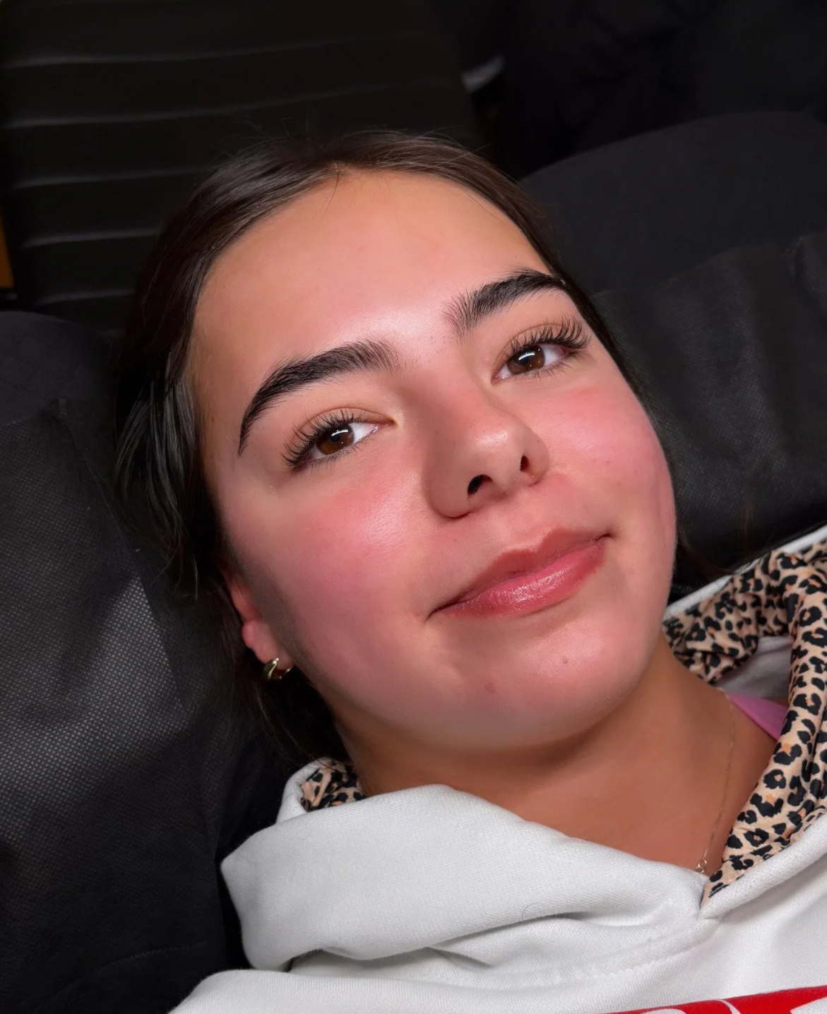 Close-up of a woman lying on a bed, looking up. She has long, curly blonde hair, pearl earrings, and is wearing a black shirt. In the background, there's a pink mug, a small wooden table, and a bed with cream-colored sheets.