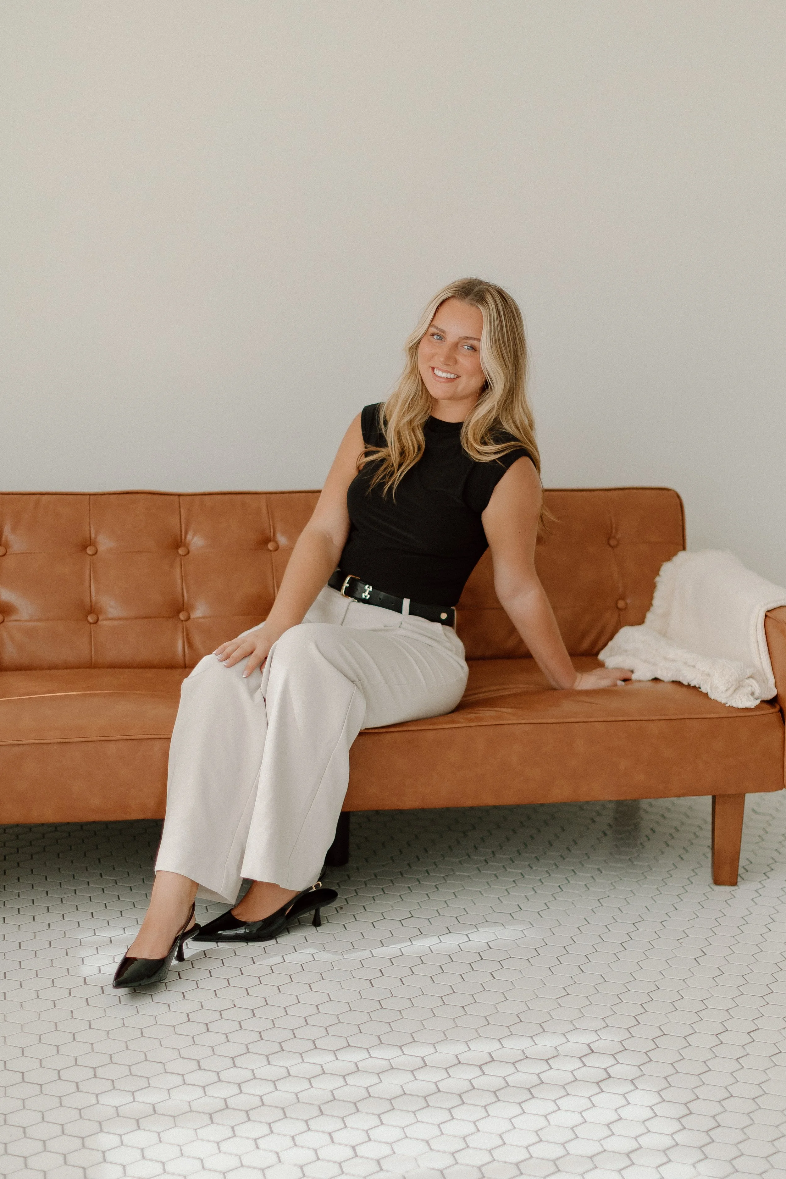 A young woman with long blonde hair, wearing a black sleeveless top, white pants, and black heels, sitting on a brown leather sofa in a room with white walls and hexagonal white floor tiles, smiling at the camera.