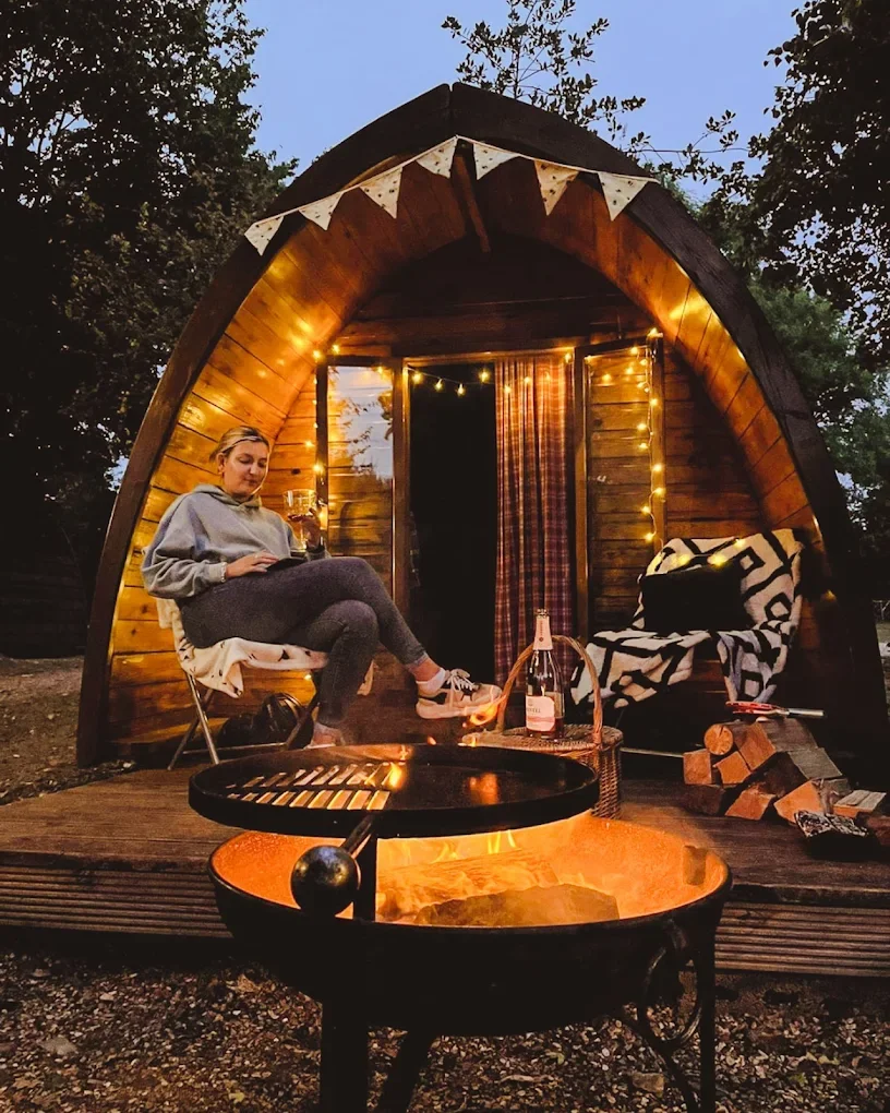 A woman sitting on a chair inside a small wooden cabin with a fire pit in front, string lights, and a cozy outdoor setting at dusk.