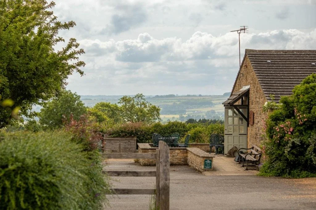 A rustic stone building with a sloped roof, surrounded by greenery and outdoor seating, overlooking a rural landscape with rolling hills and cloudy sky.