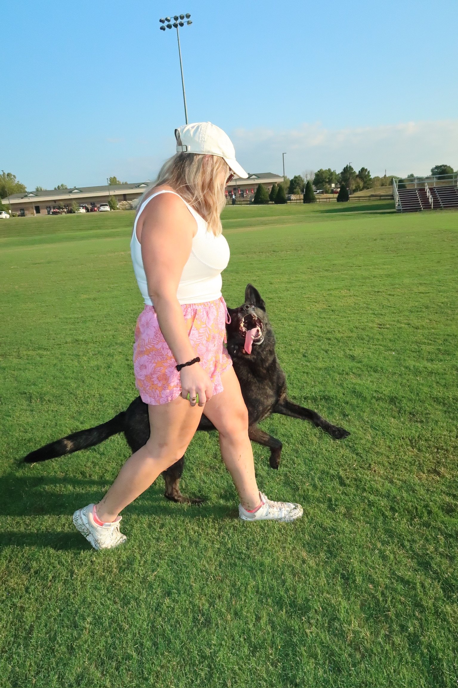 A woman in a white tank top, pink floral shorts, and white sneakers is running on a grassy field with her black dog, which has its tongue out and ears perked up.