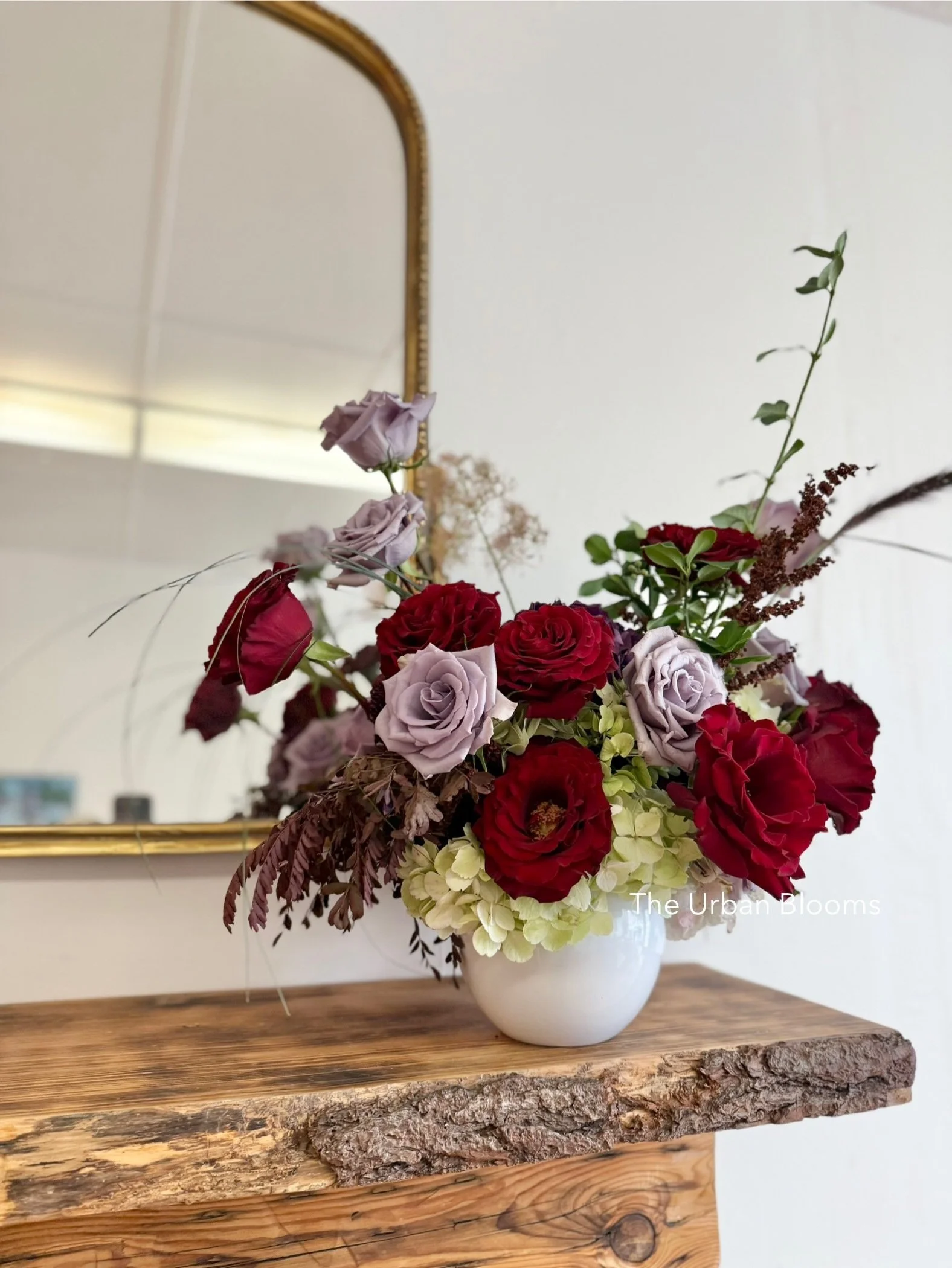 A floral arrangement with red, light purple, and cream flowers in a white ceramic vase on a wooden table.
