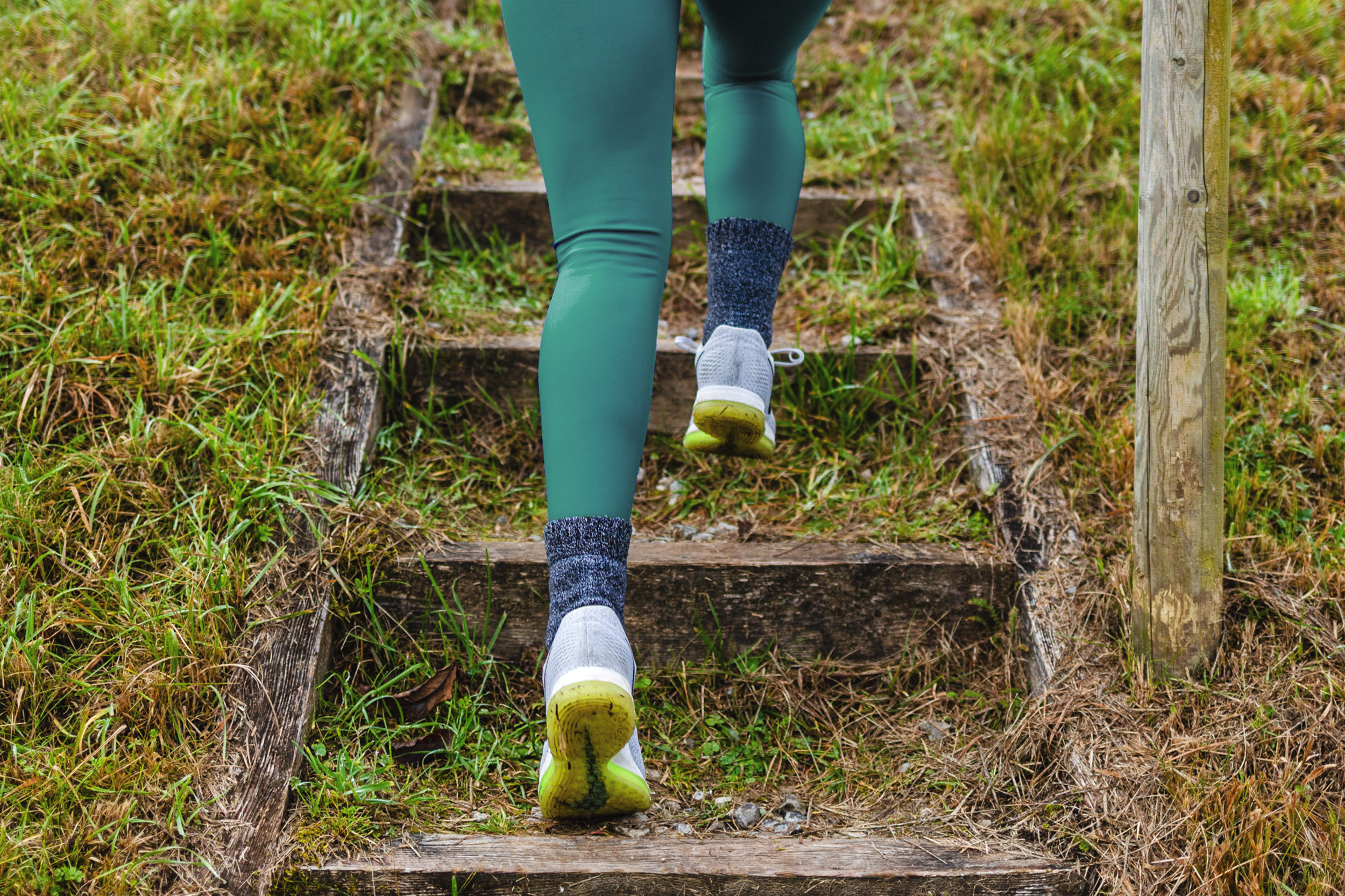 Person, die auf einer Holztreppe im Freien bergauf geht, trägt Sportschuhe, Leggings und dicke Socken, umgeben von Gras.