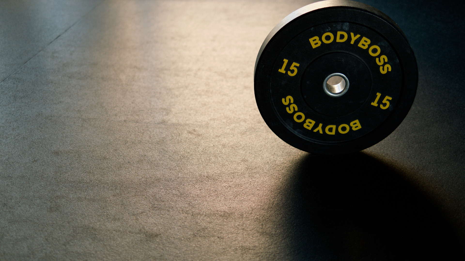A black dumbbell weight plate with yellow lettering that reads 'BODYBOSS 15' is on a dark textured gym floor, casting a shadow to the right.