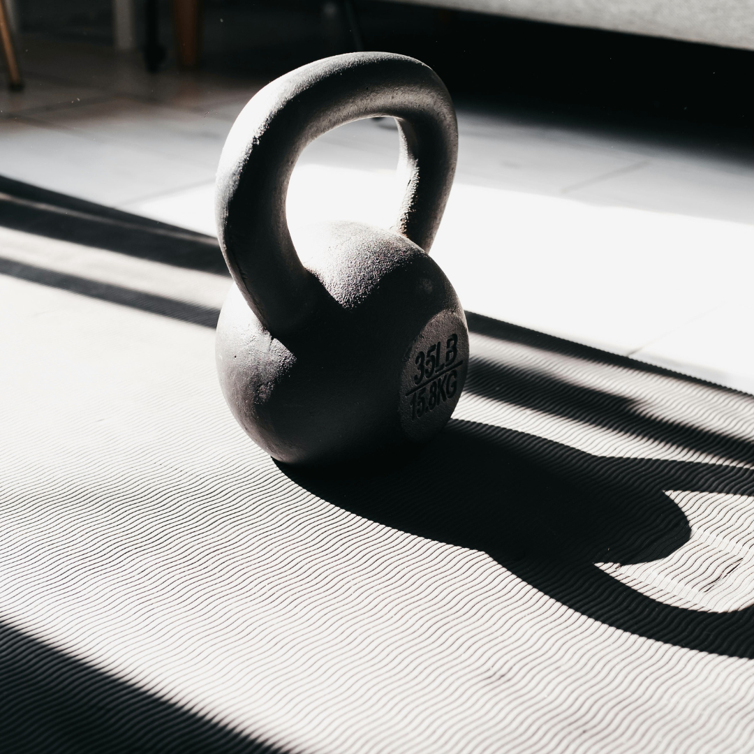 A black kettlebell with a handle, casting a shadow on a textured gym mat in sunlight.