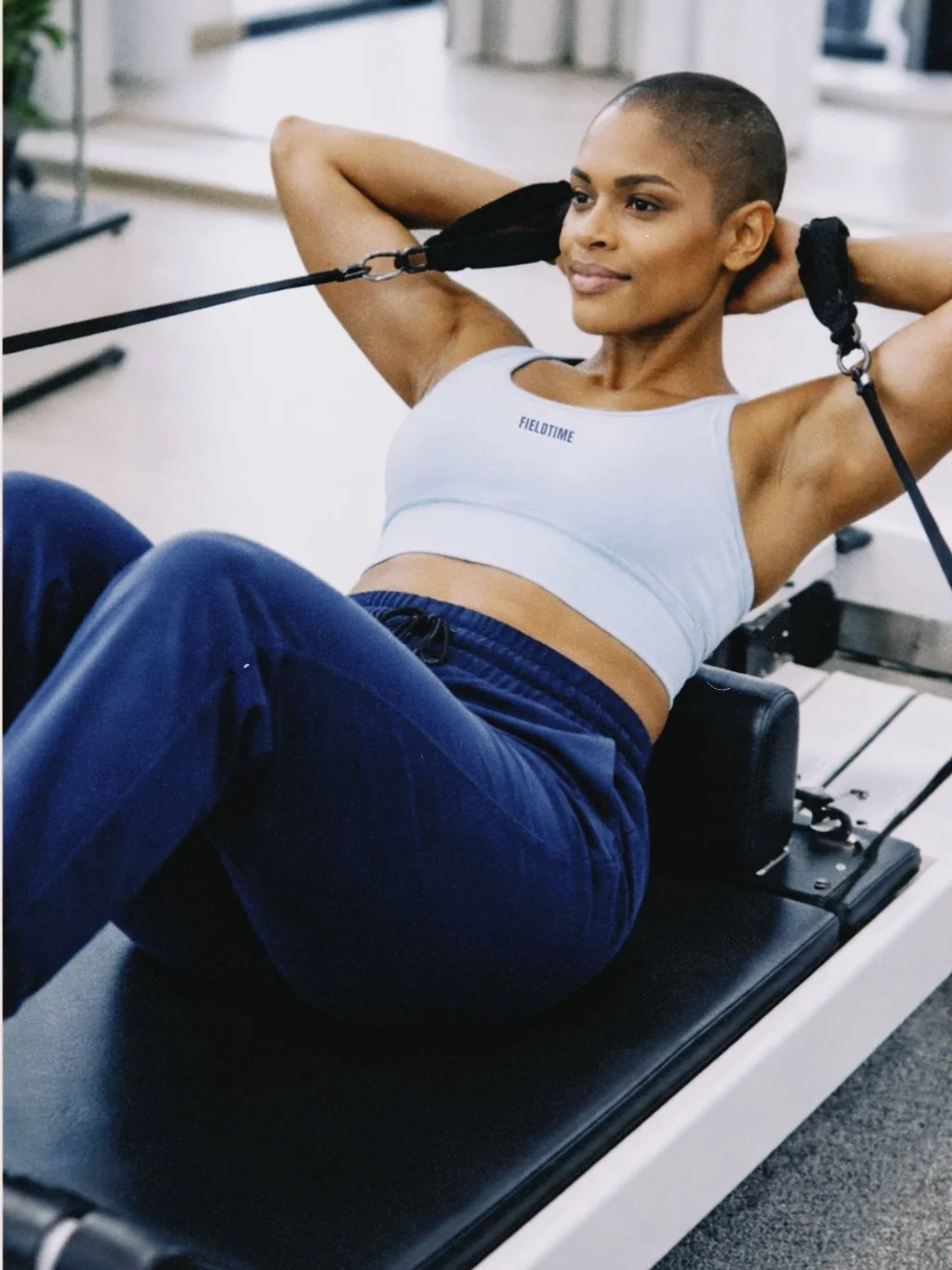 A woman with a buzz cut hair wearing a white sports bra and navy blue pants, performing sit-ups with resistance straps in a gym.