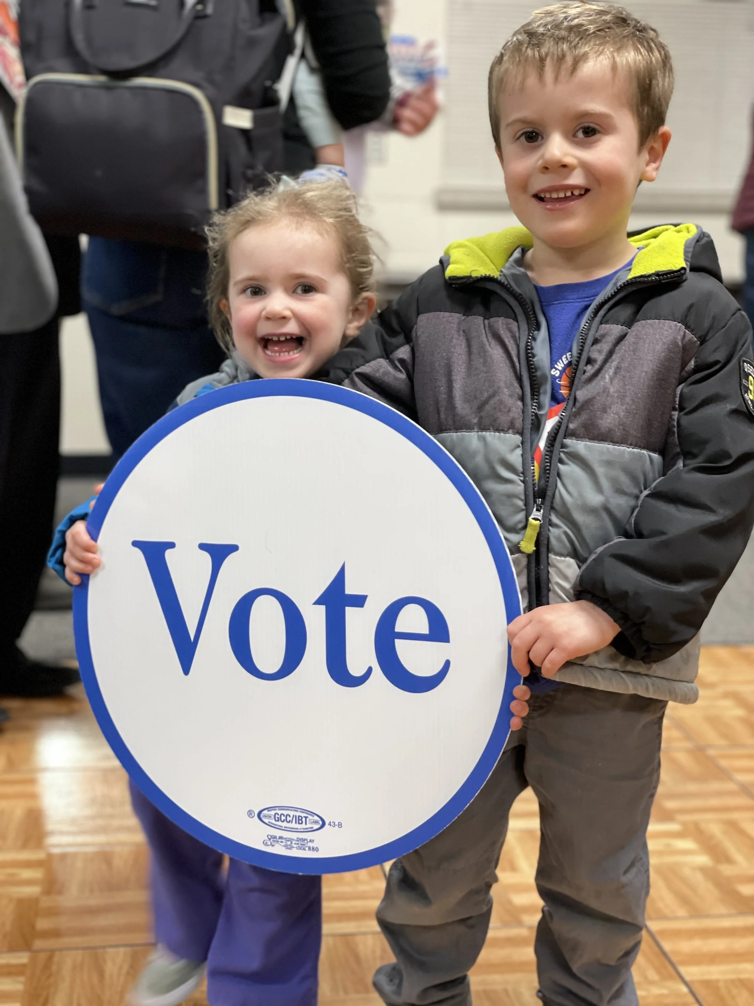 Christos's children, a boy and a girl, holding a large white sign with blue border and the word 'Vote' written on it. The boy is wearing a black and gray jacket and smiling. The girl is standing behind the sign, wearing a purple outfit, .