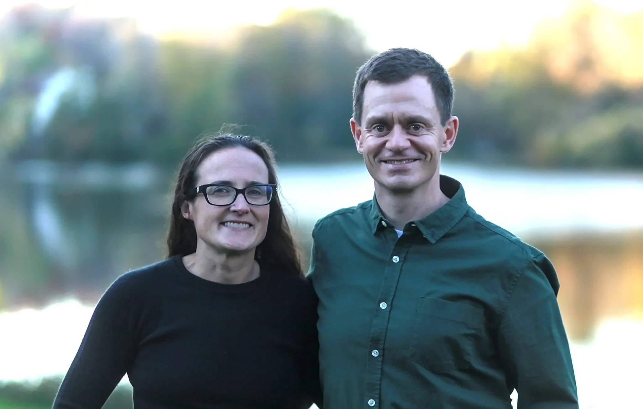 Christos and his wife standing outdoors near a body of water with trees in the background.