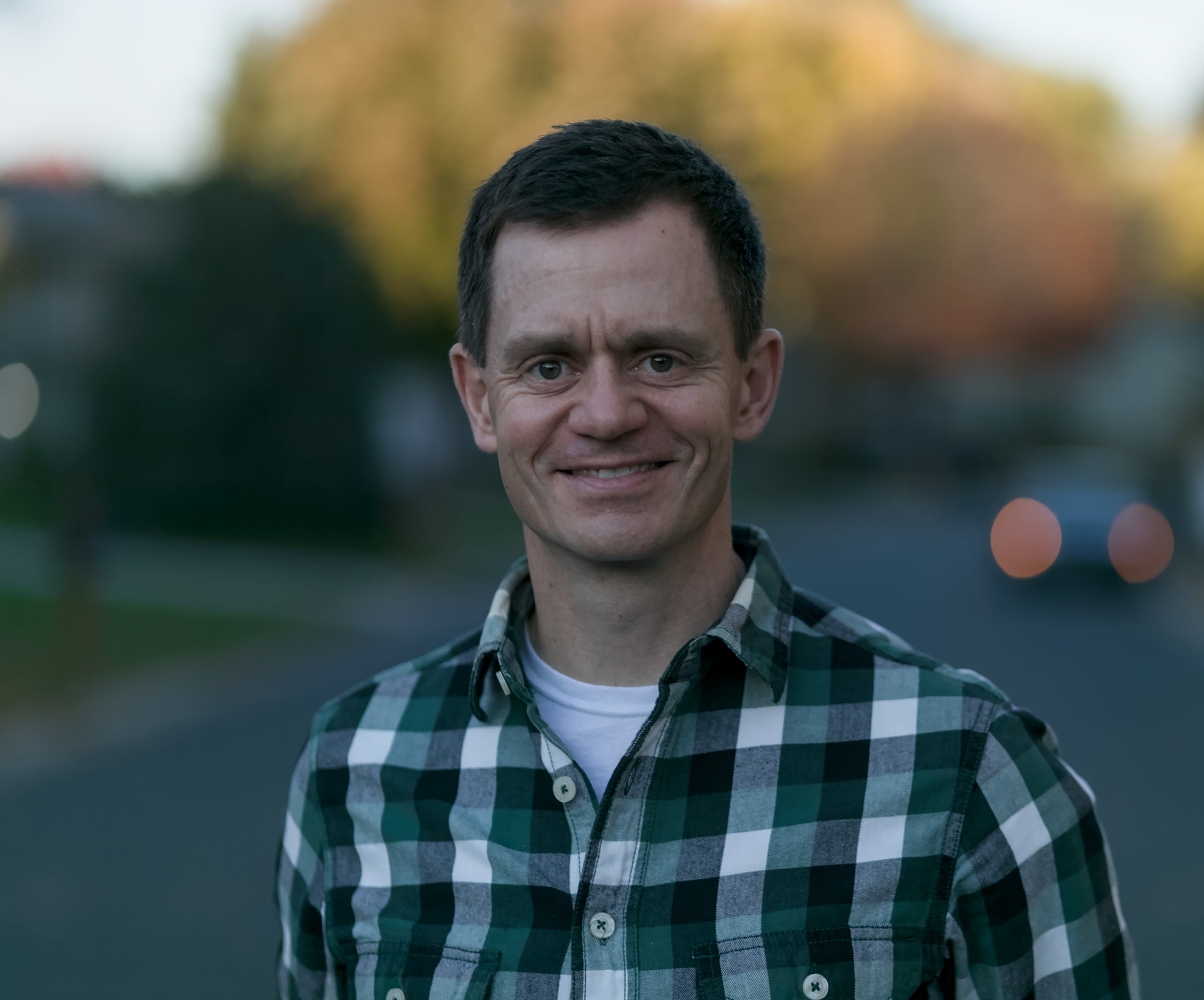 Christos wearing a green and black plaid shirt, standing outdoors with blurred trees and street in the background during sunset.