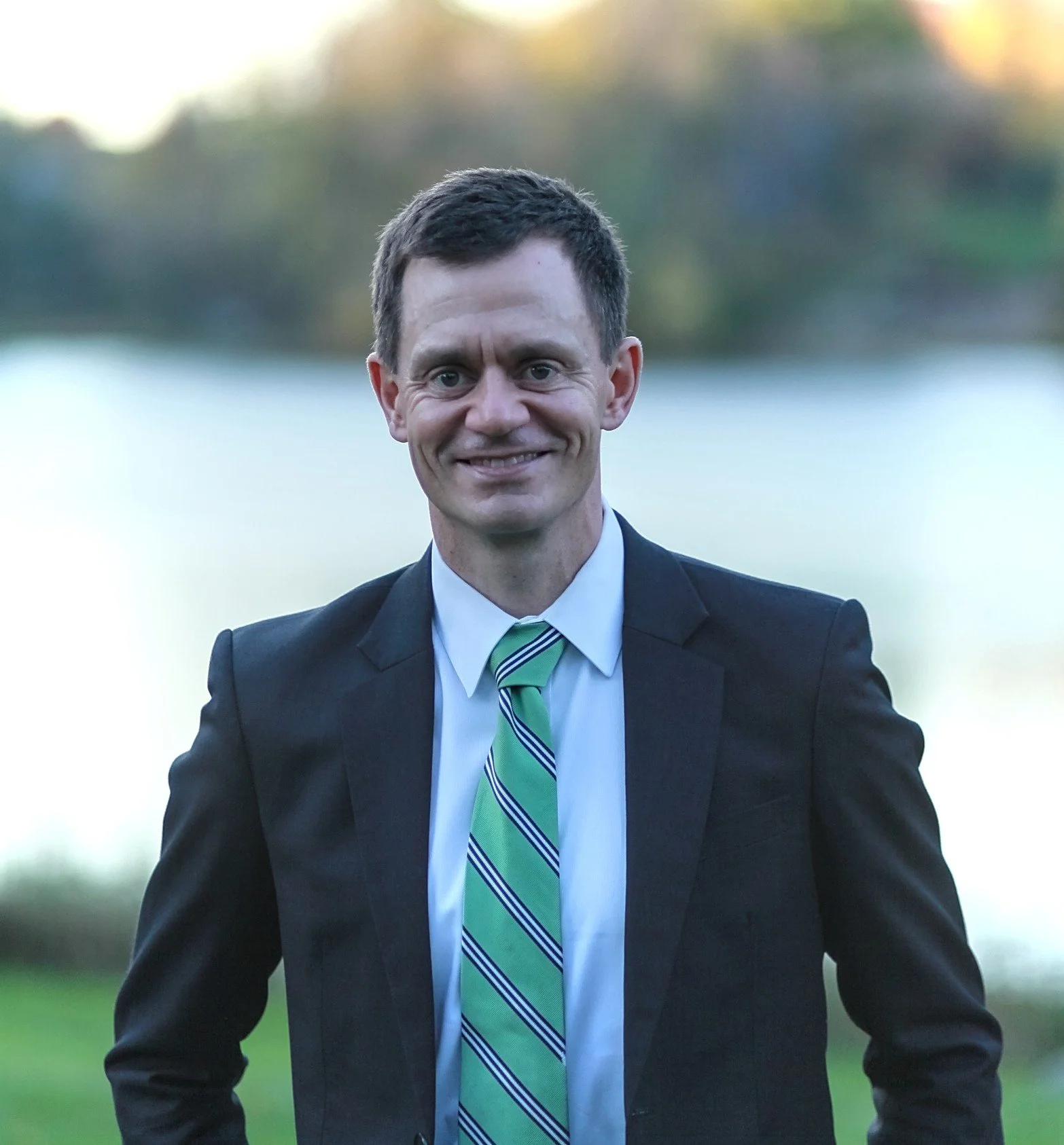 Christos in a suit with a green striped tie standing outdoors near a body of water.