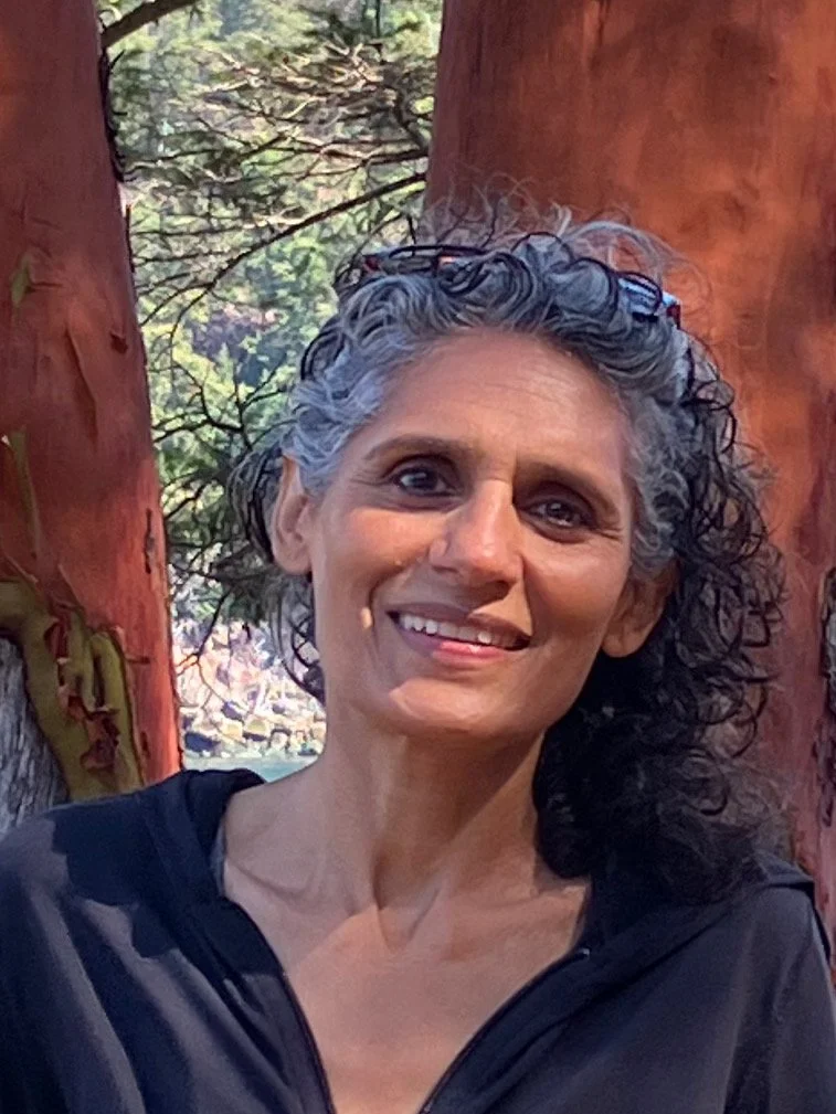A woman with short curly gray hair smiling in front of a red tree trunk and green forest in the background.