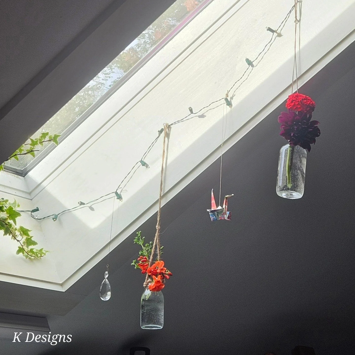 Hanging decorations from a skylight window, including glass jars with flowers and an origami crane, with string lights and green plants nearby.
