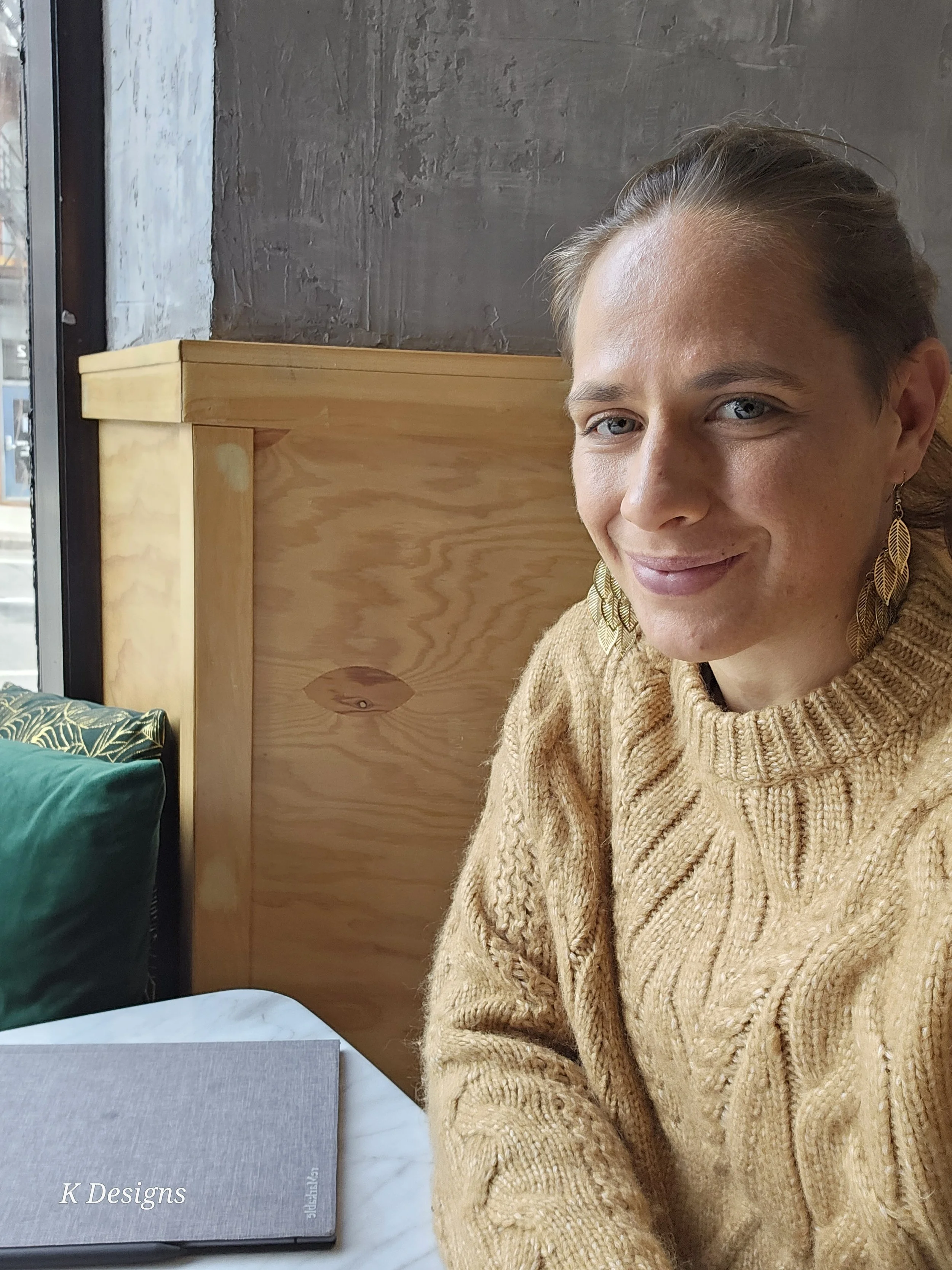 A woman with blonde hair wearing large leaf-shaped earrings and a tan cable-knit sweater sitting at a table in a cafe, smiling at the camera. There is a window to her left, a wooden partition behind her, and a gray menu on the table.