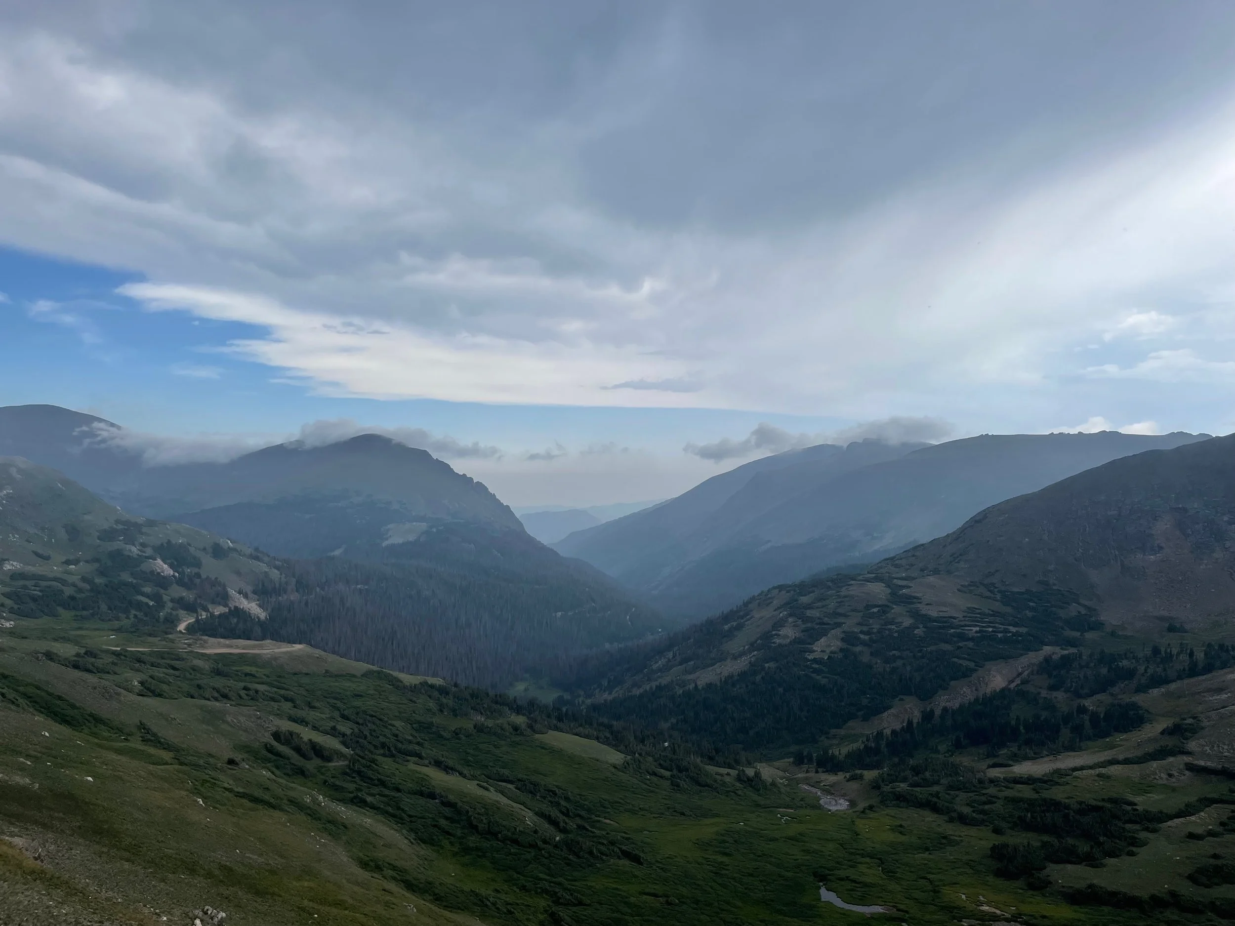 A scenic view of a mountainous landscape with lush green valleys, rolling hills, and distant mountains under a mostly cloudy sky.