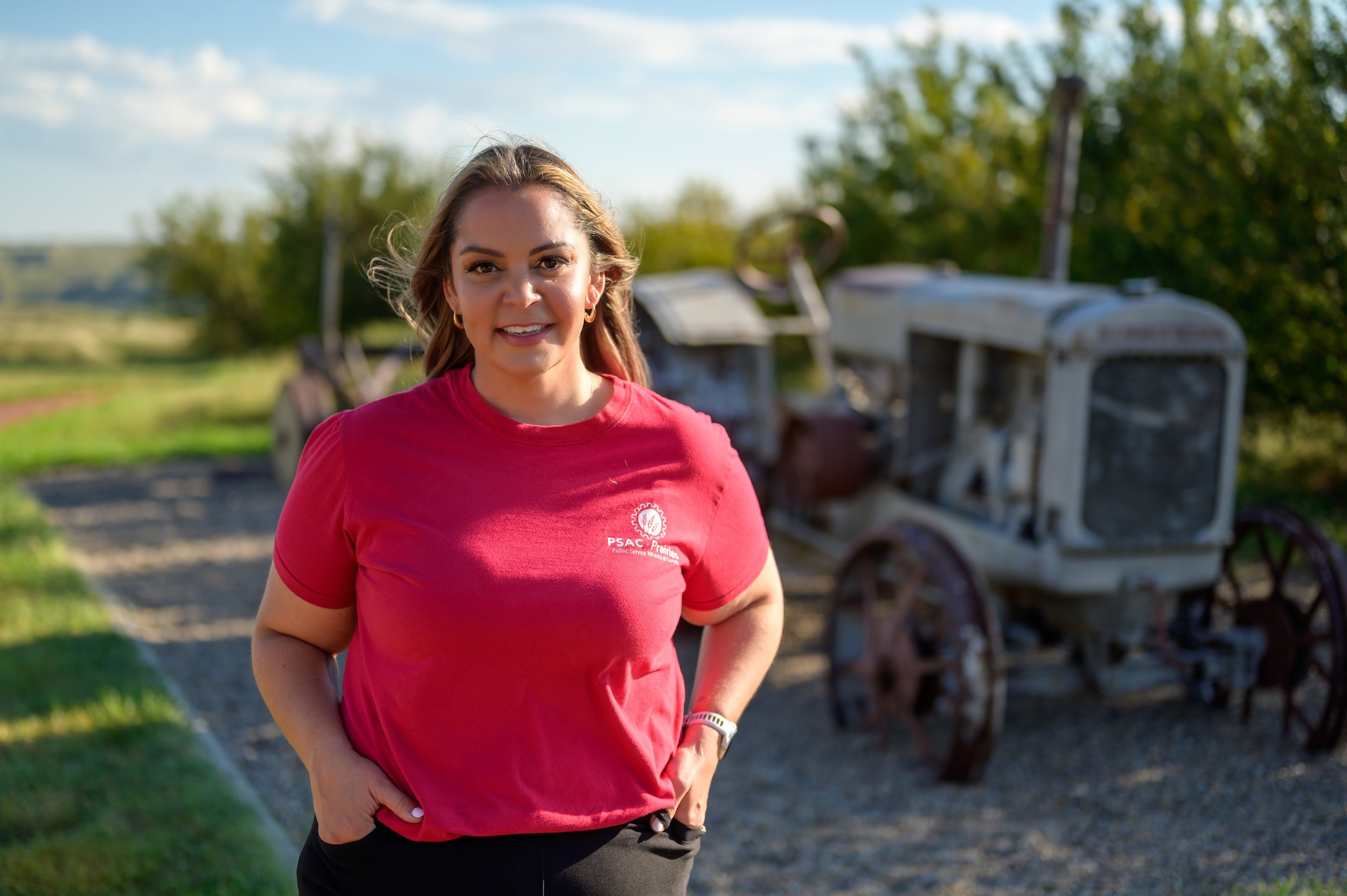 A woman in a red t-shirt standing outdoors with an old tractor and greenery in the background.