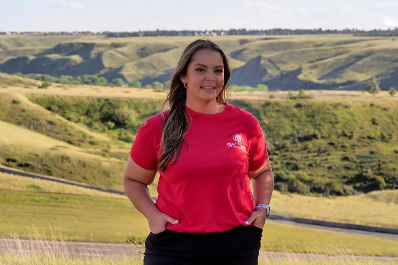 Woman standing outdoors with rolling green hills in the background, wearing a red t-shirt and black pants.