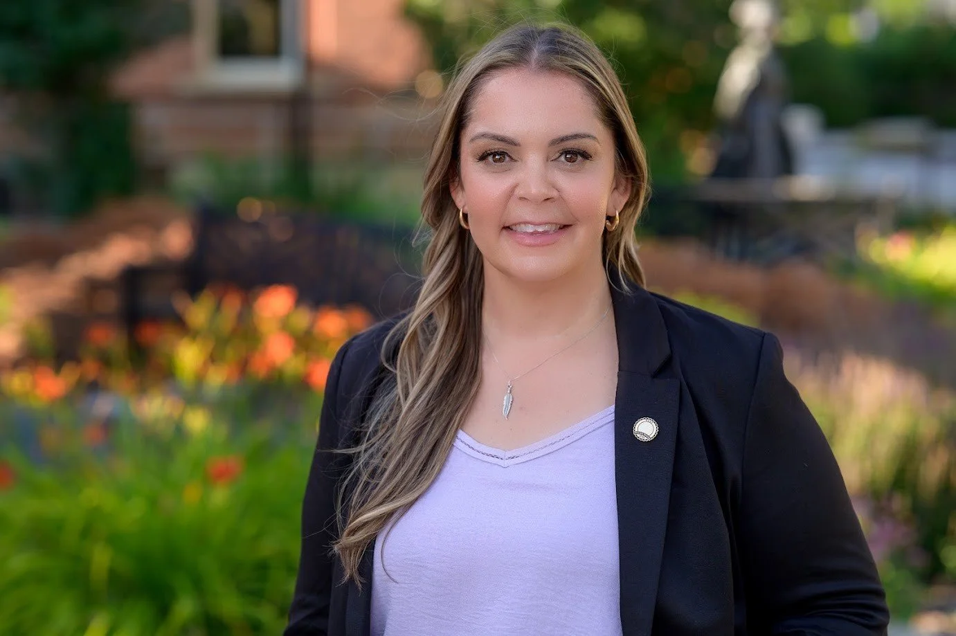 Portrait of a woman with light brown hair wearing a black blazer, white shirt, and jewelry, standing outdoors in a garden.