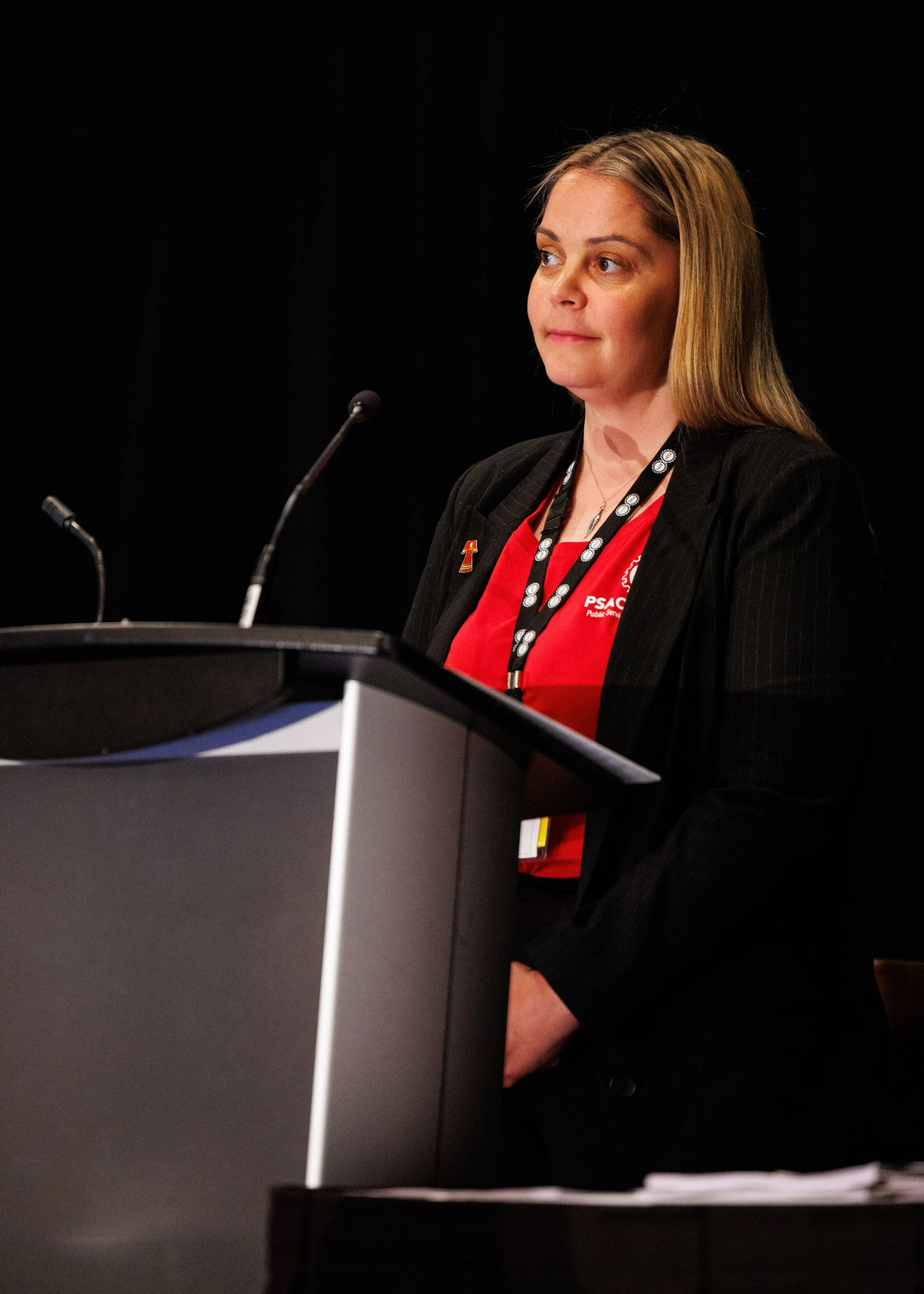 A woman standing at a podium, wearing a black blazer and red shirt with PSL, speaking at a conference or event.