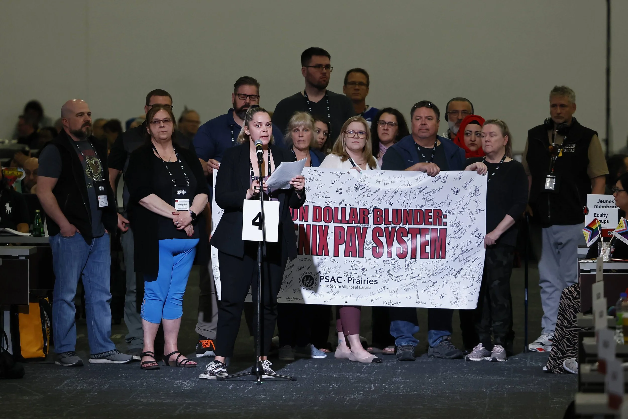 Krysty Thomas speaks at a rally with a group of people holding a sign that reads 'One Billion Dollar Blunder: Phoenix Pay System' with signatures.