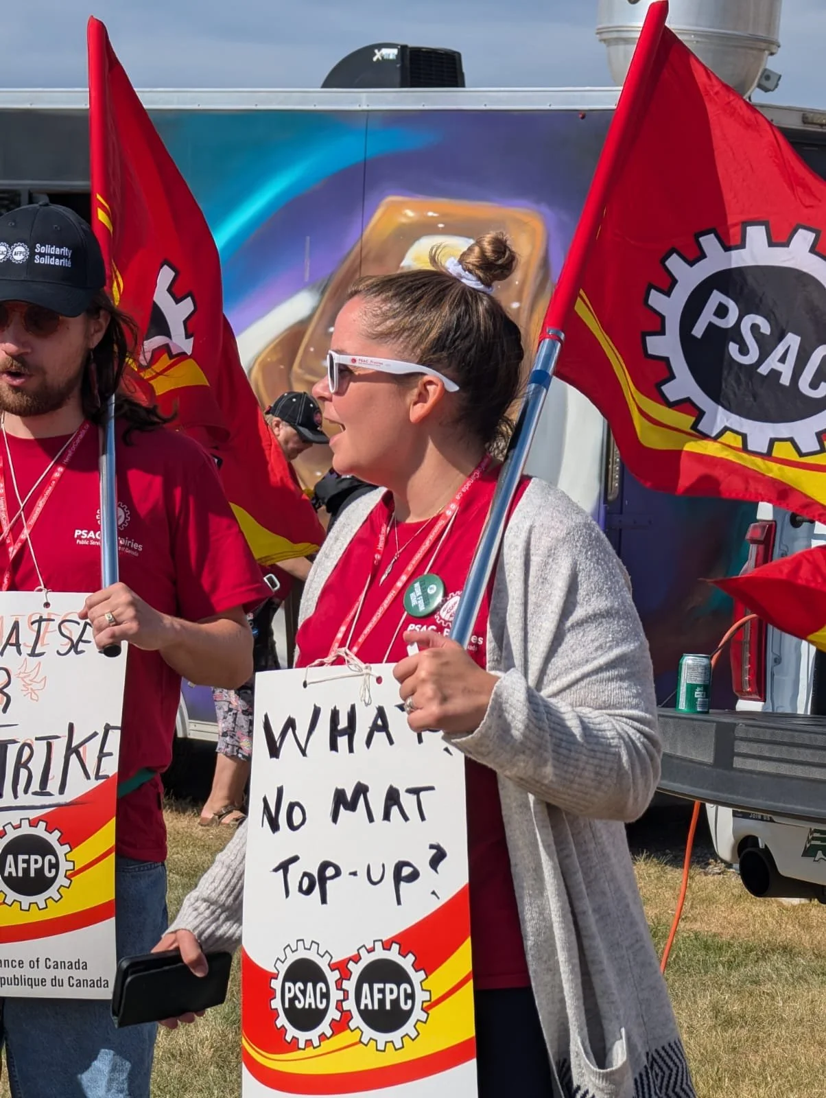 Krysty Thomas participating in a rally, holding signs and flags, with one sign reading "WHAT NO MAT TOP-UP?" and flags displaying the PSAC logo. They are outdoors, and some individuals are wearing red shirts.