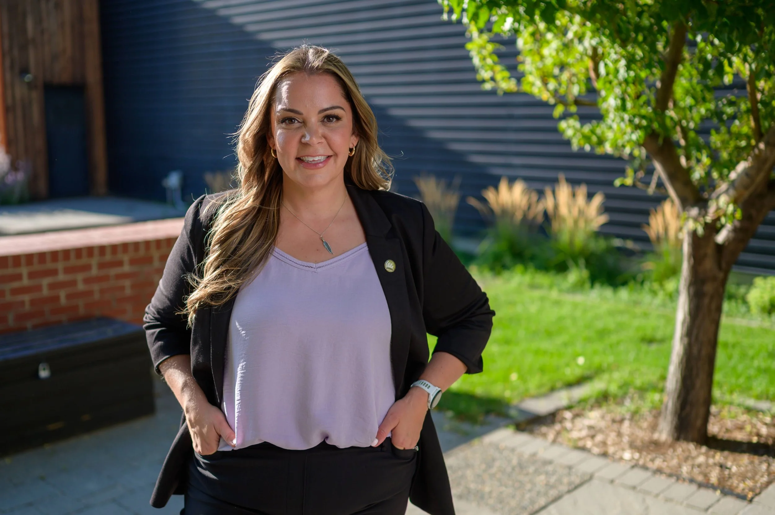 A woman with long, wavy blonde hair smiling outdoors, wearing a black blazer over a light-colored top, standing in a garden with green grass, trees, and a brick and metal fence.