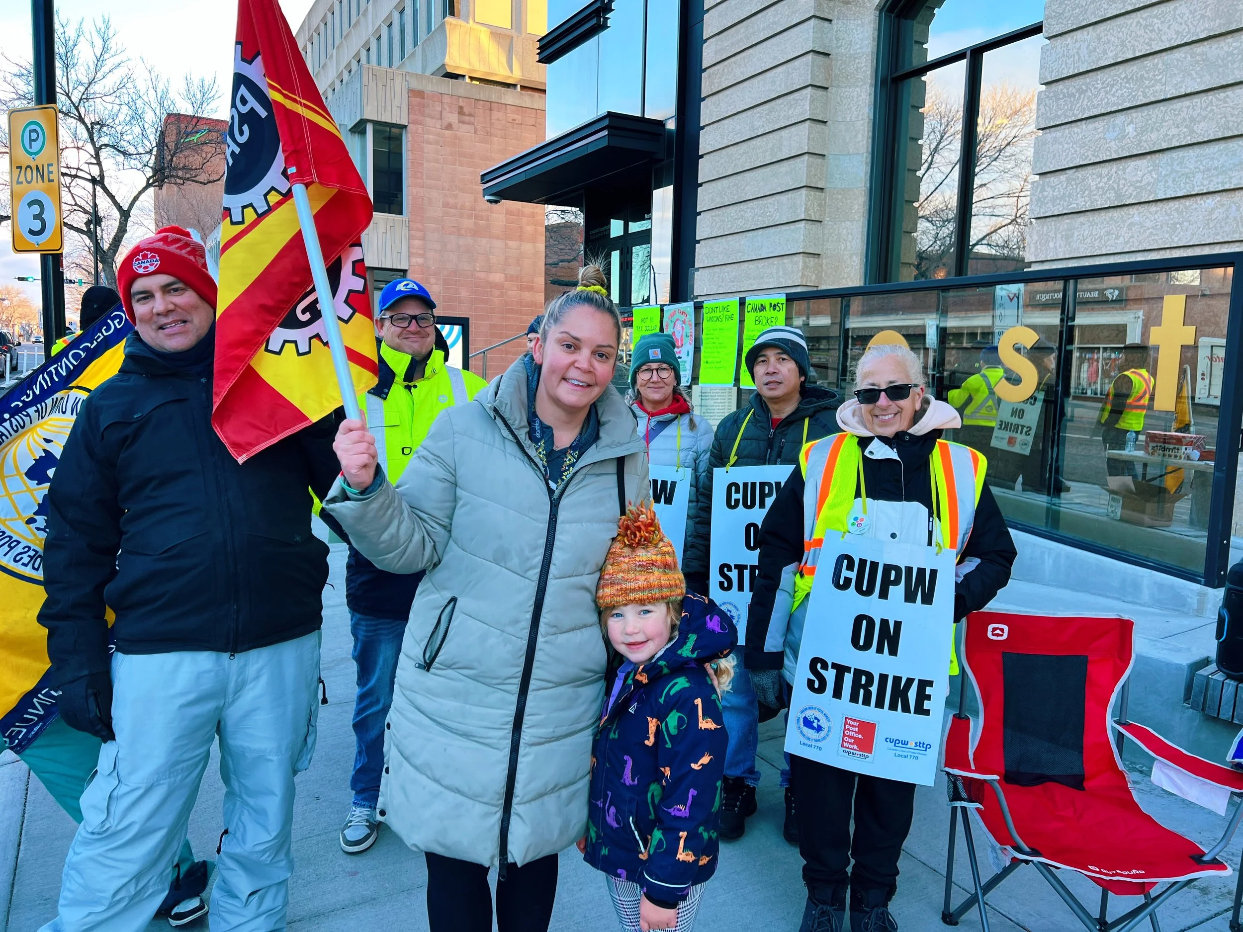 Krysty Thomas stands at a protest holding flags and signs that read “CUPW ON STRIKE,” showing support for postal workers.