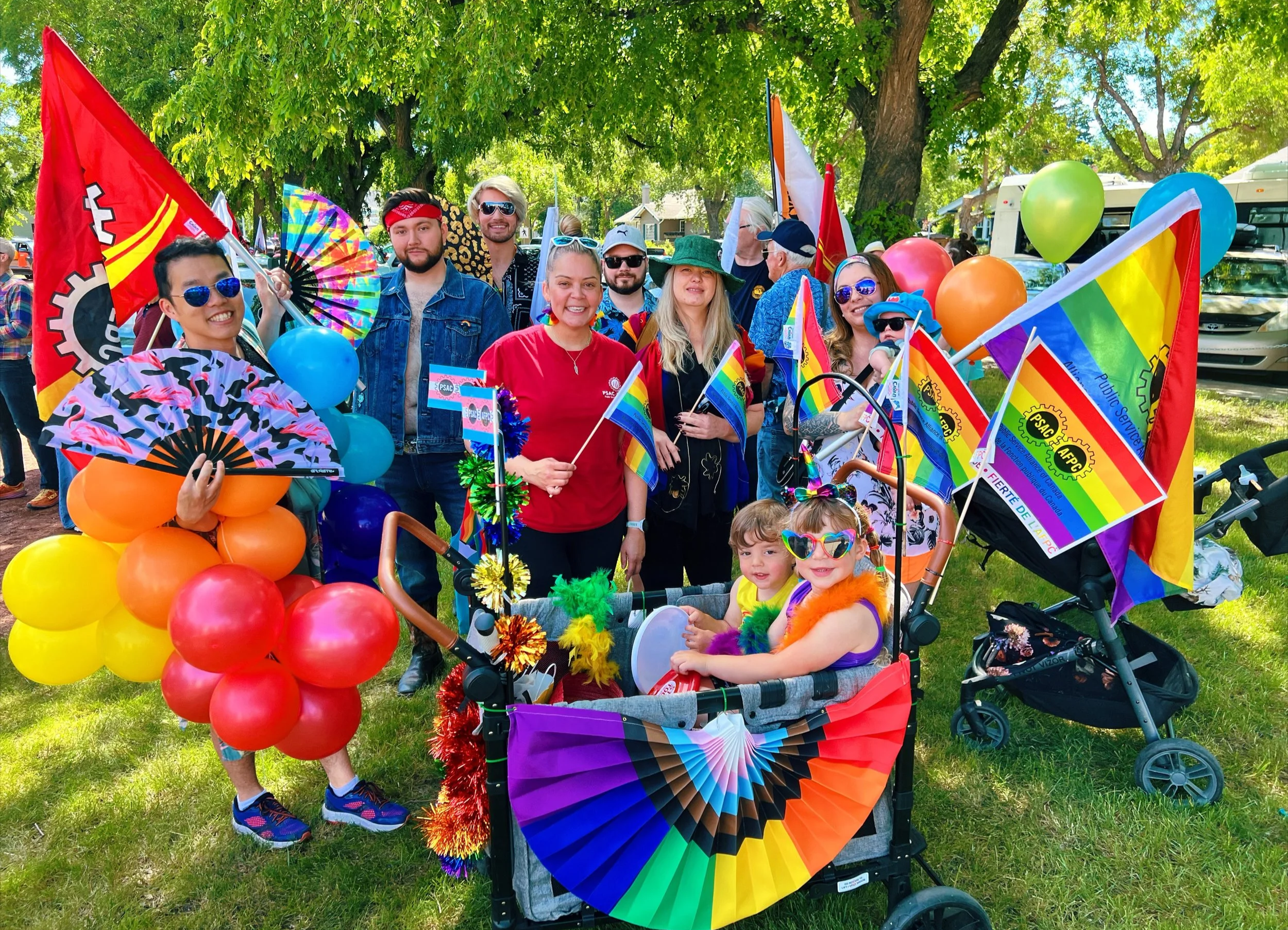 Group of people celebrating at a pride parade with rainbow flags, balloons, fans, and vibrant clothing, including children in a decorated stroller.