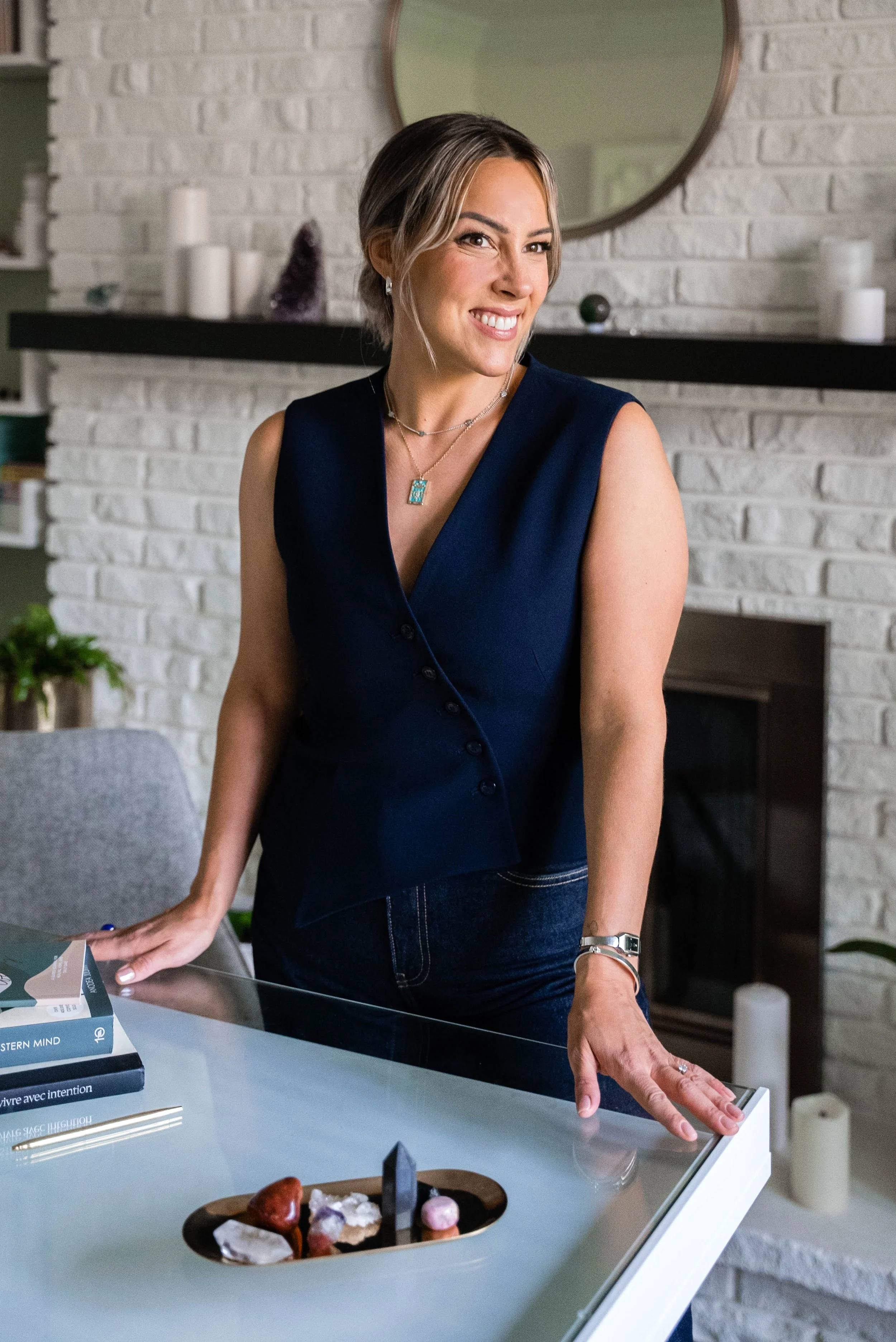 A woman with blonde hair smiling and leaning on a glass table in a living room with a white brick fireplace and decorated mantle.