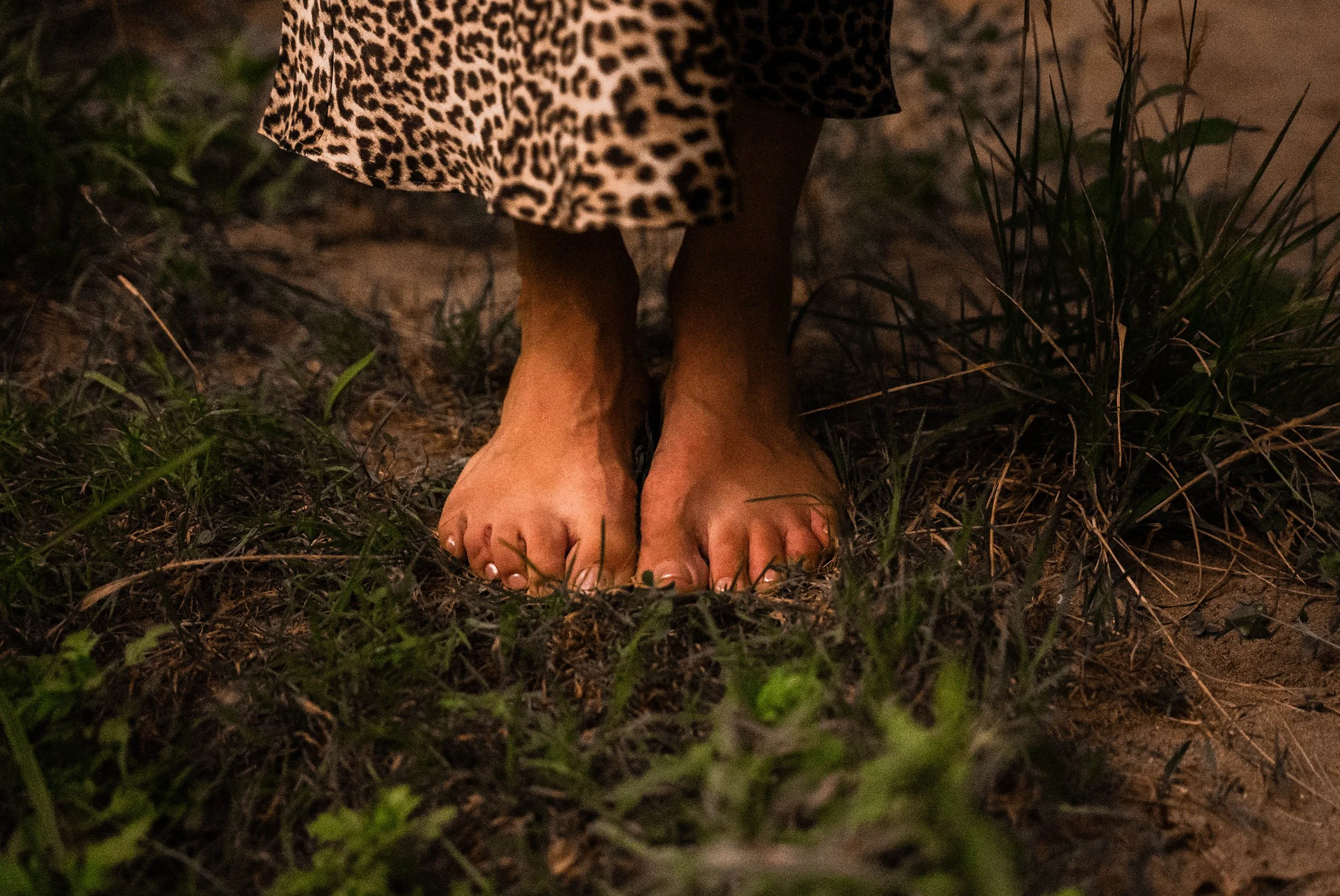 Close-up of a person's bare feet standing on a dirt ground surrounded by grass, with part of a leopard print dress visible.