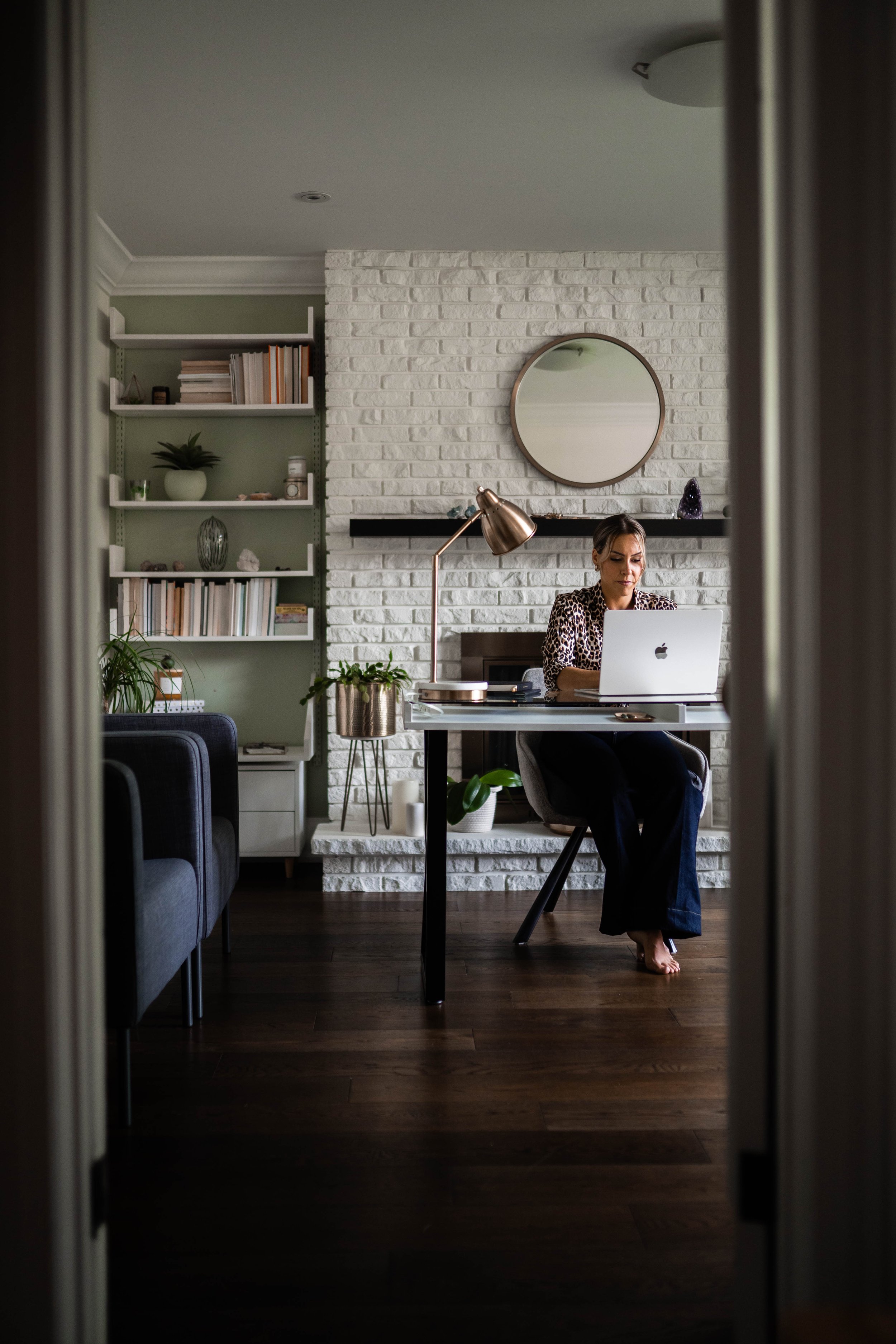 A woman working on a laptop at a desk in a living room with white brick fireplace, round mirror, bookshelf, and plants.