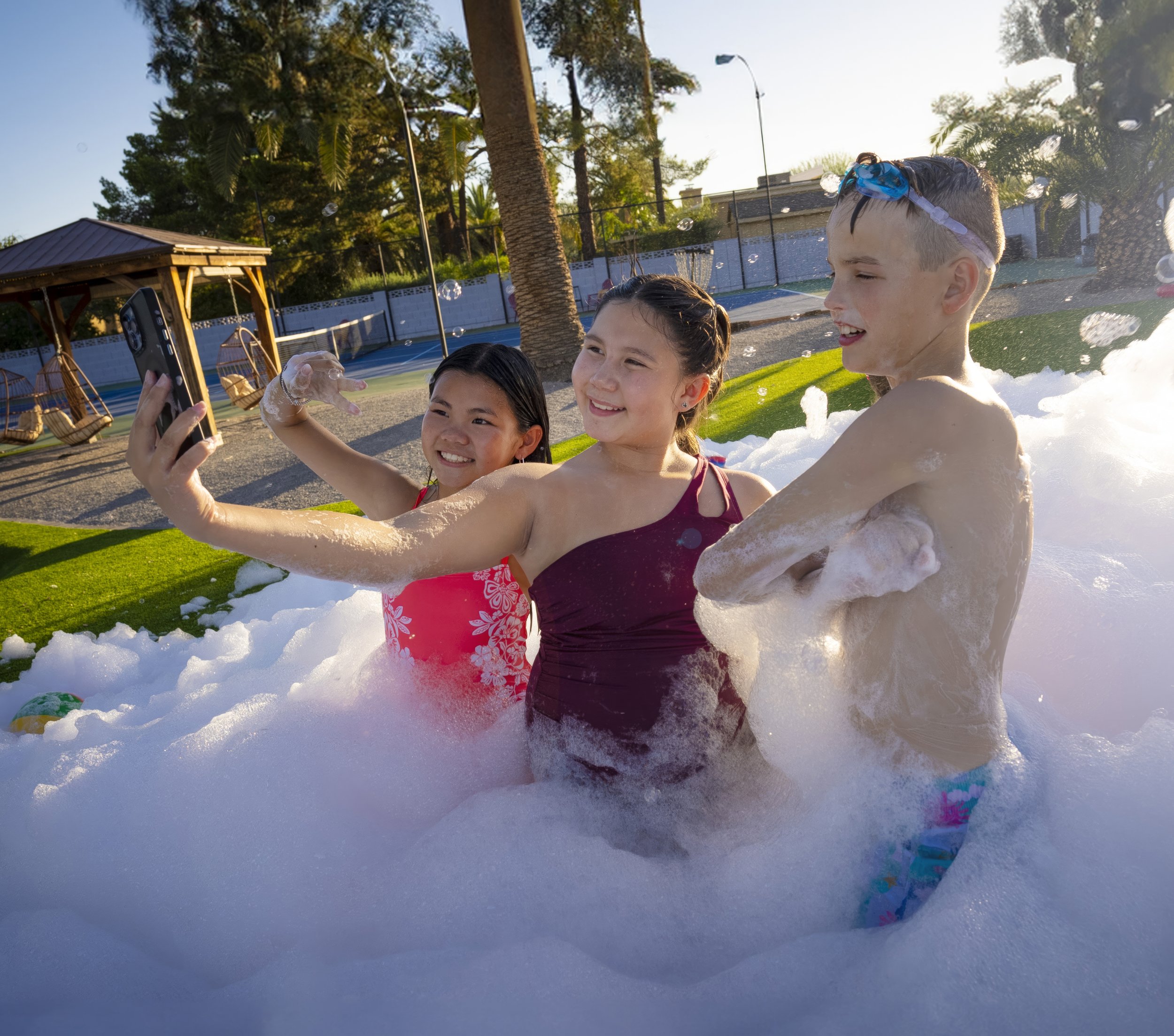 Three children in a foam-filled pool taking a selfie, smiling and enjoying a sunny day.